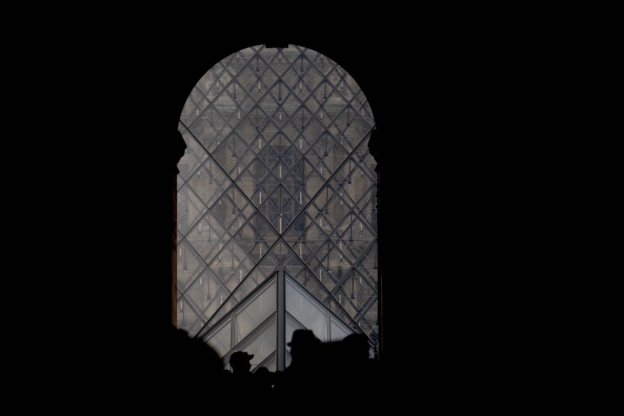 写真は、フランスのパリにあるルーブル美術館のピラミッドを捉えています。ピラミッドは、暗いアーチ状の開口部を通して見えています。アーチの黒いシルエットが、ピラミッドの幾何学的な構造を際立たせています。ピラミッドのガラスと金属のフレームが作り出す模様は複雑で、光の反射によってさらに強調されています。アーチの下部には、人々のシルエットがわずかに見え、ピラミッドの壮大さと人気を示唆しています。全体的に、写真はコントラストが強く、神秘的で、建築的な美しさを強調しています。
