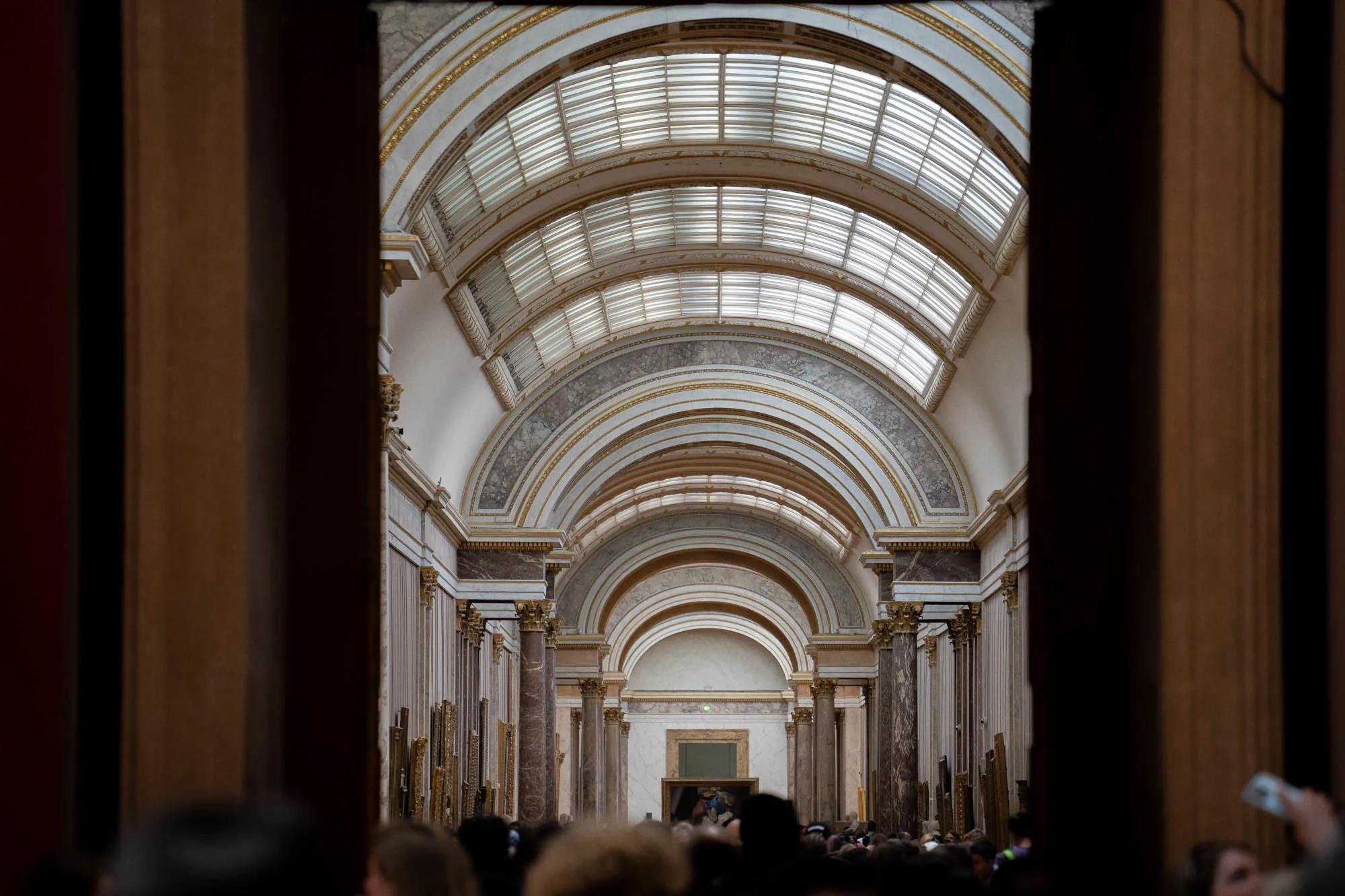 写真はフランスのパリにある美術館の内部を捉えたものです。カメラは、木の柱で縁取られた開口部から、奥へと続くアーチ状の天井を持つ長いギャラリーを覗き込んでいます。天井は、曲線を描くリブと格子状の窓で構成され、自然光を取り込んでいます。ギャラリーの両側には、豪華な大理石の柱が立ち並び、壁には金色の額縁に入れられた絵画が多数掛けられています。ギャラリーの奥には、さらにアーチが続き、その先に人が集まっている様子が見えます。写真は、壮麗で歴史的な雰囲気を醸し出し、芸術と文化の中心地としてのパリの魅力を伝えています。