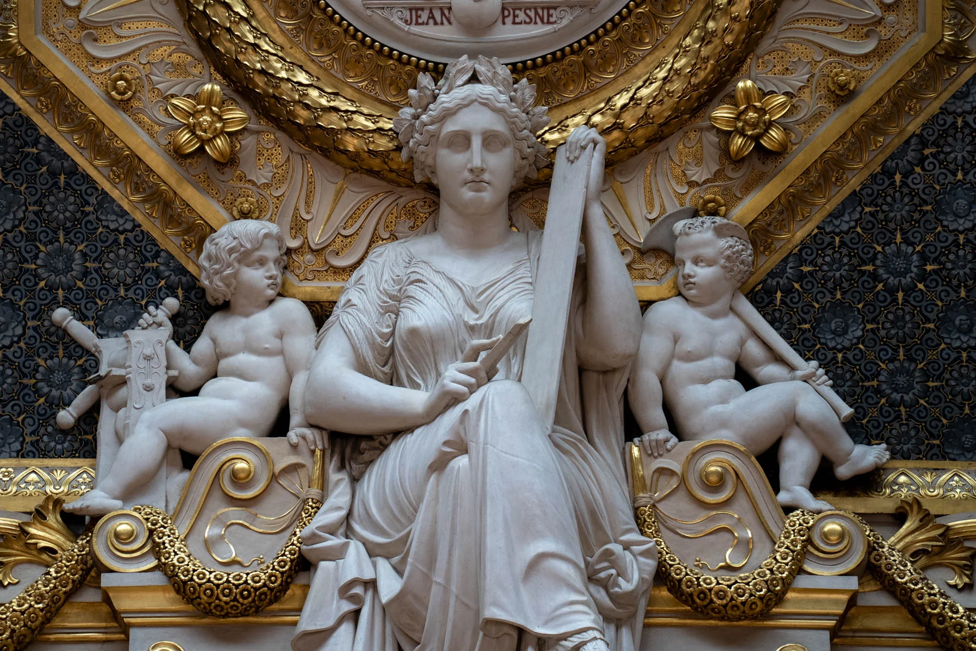 写真には、フランスのパリにあると思われる、精巧な装飾が施された彫刻が写っています。中央には、古代ギリシャ風の衣装を身につけた女性の白い彫像が座っています。彼女は頭に月桂樹の冠を戴き、左手に何かを書き記すための石板のようなものを持っています。彼女の左右には、それぞれキューピッドのような愛らしい天使の彫像が配置されており、それぞれが何らかの道具を持っています。背景には、金色の装飾や花柄の装飾が施されており、非常に豪華で荘厳な雰囲気を醸し出しています。像の上部には「JEAN PESNE」という文字が見えます。全体的に、古典的な美しさと豪華さが感じられる写真です。