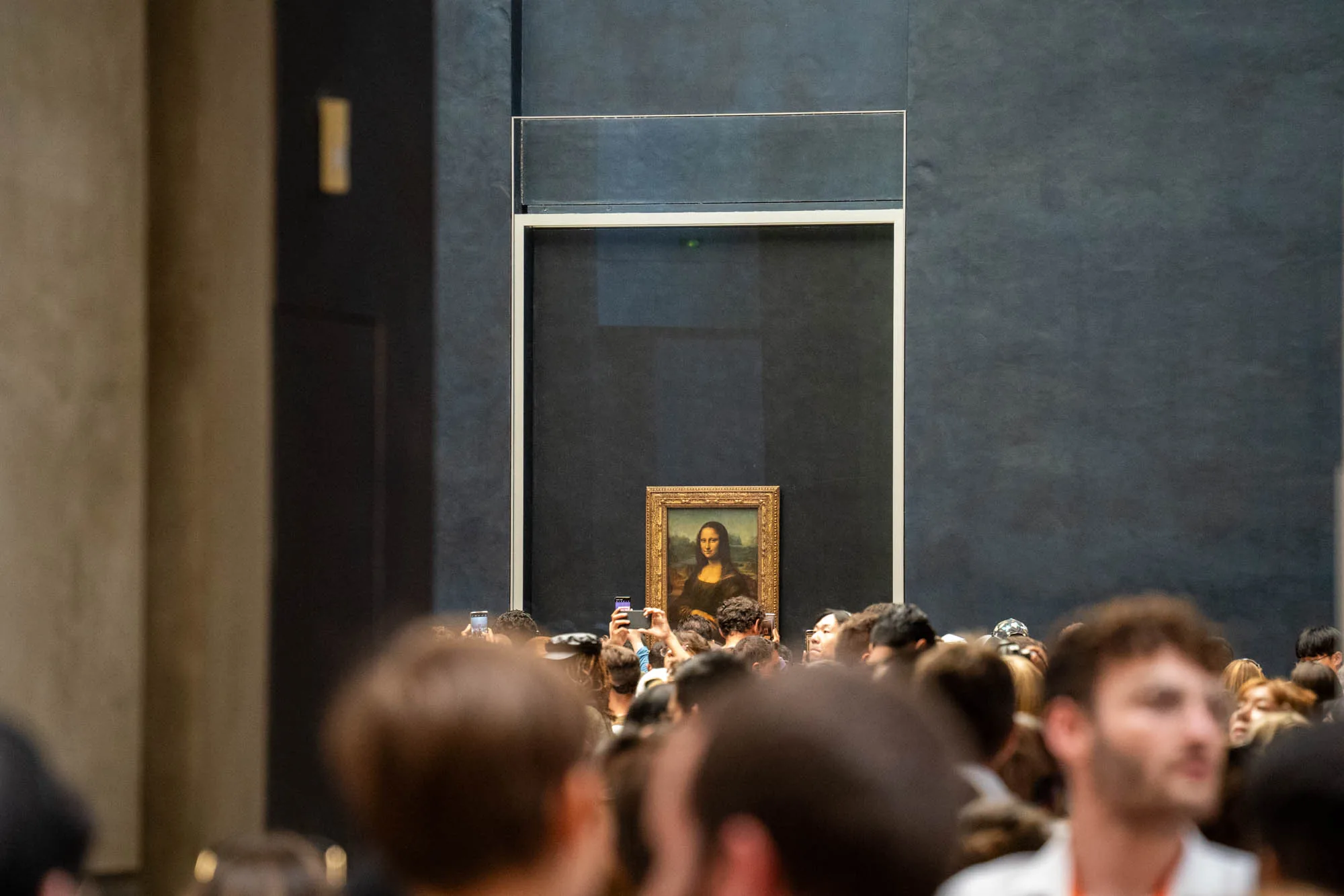 この写真は、フランスのパリにあるルーブル美術館で撮影されたものでしょう。画面奥には、金色の額縁に入ったレオナルド・ダ・ヴィンチの有名な絵画「モナリザ」が見えます。絵画は、ダークグレーの壁に展示されています。  絵画の前には、大勢の人が集まっており、絵画を鑑賞したり、スマートフォンで写真を撮ったりしています。多くの人の頭部が写っており、鑑賞者の熱気と混雑ぶりを伝えています。人々の顔はぼやけていますが、その視線は一様にモナリザに向けられているようです。  絵画の上部には、ガラス製の保護ケースのようなものが見え、絵画が厳重に保護されていることがわかります。全体的に、名画をひと目見ようと集まった人々の熱気と、その作品を保護するための厳重な措置が伝わってくる写真です。