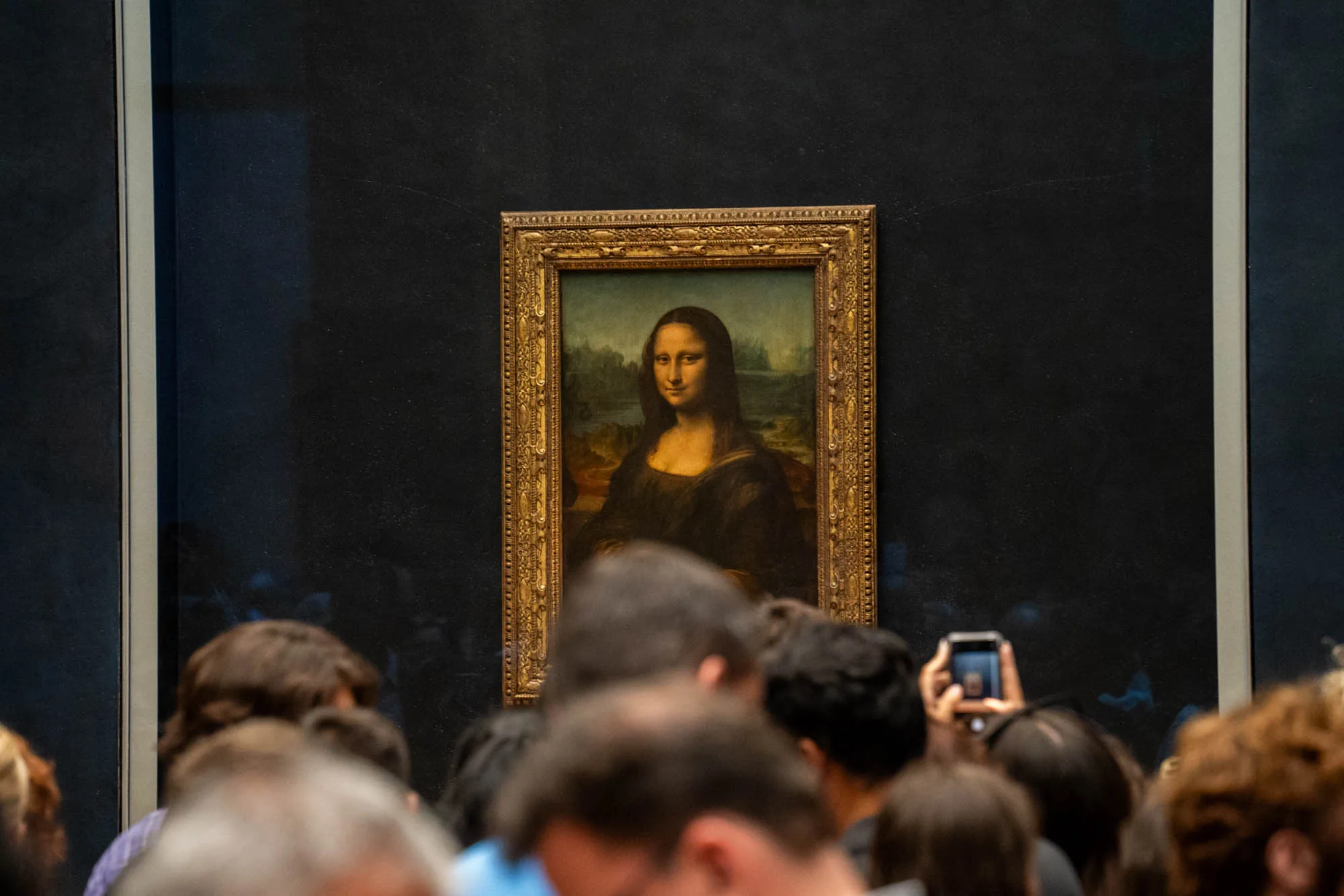 写真は、フランス・パリのルーブル美術館で展示されているレオナルド・ダ・ヴィンチの傑作「モナリザ」を捉えたものです。絵画は、豪華な金色の額縁に飾られ、濃い青色の壁にかけられています。絵画の前には大勢の観客がおり、その頭部が前景に写っています。人々はスマートフォンなどで写真を撮っており、その様子から人気の高さが伺えます。作品を鑑賞しようと集まった人々の熱気が伝わってくるような雰囲気です。