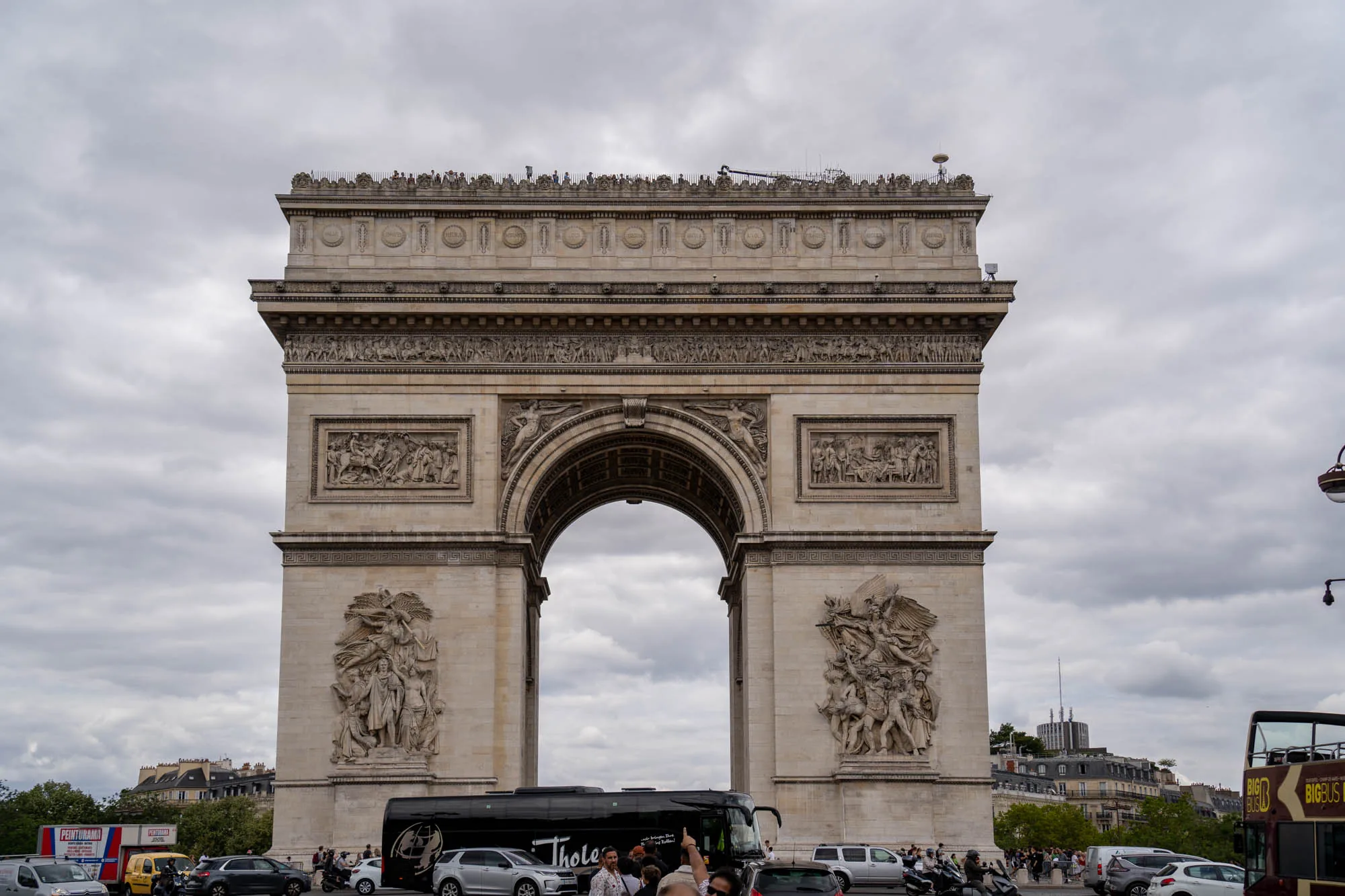 写真には、フランスのパリにある有名な建造物、凱旋門が写っています。空は曇り空で、重たい雲が広がっています。凱旋門は、壮大で堂々とした佇まいで、その表面には精巧な彫刻が施されています。門の上部には、さらに細かい装飾や彫像が並んでおり、歴史と芸術の重みを感じさせます。  凱旋門の下には、道路があり、様々な車やバスが通行しています。観光客らしき人々も行き交っており、活気があります。手前には観光バスが停車しており、観光地ならではの賑わいを醸し出しています。遠景には、パリの街並みが広がり、屋根の色合いや建物の形状から、ヨーロッパの都市らしい景観が伺えます。全体的に落ち着いた色調で、歴史的な建造物と現代の都市生活が調和した風景です。
