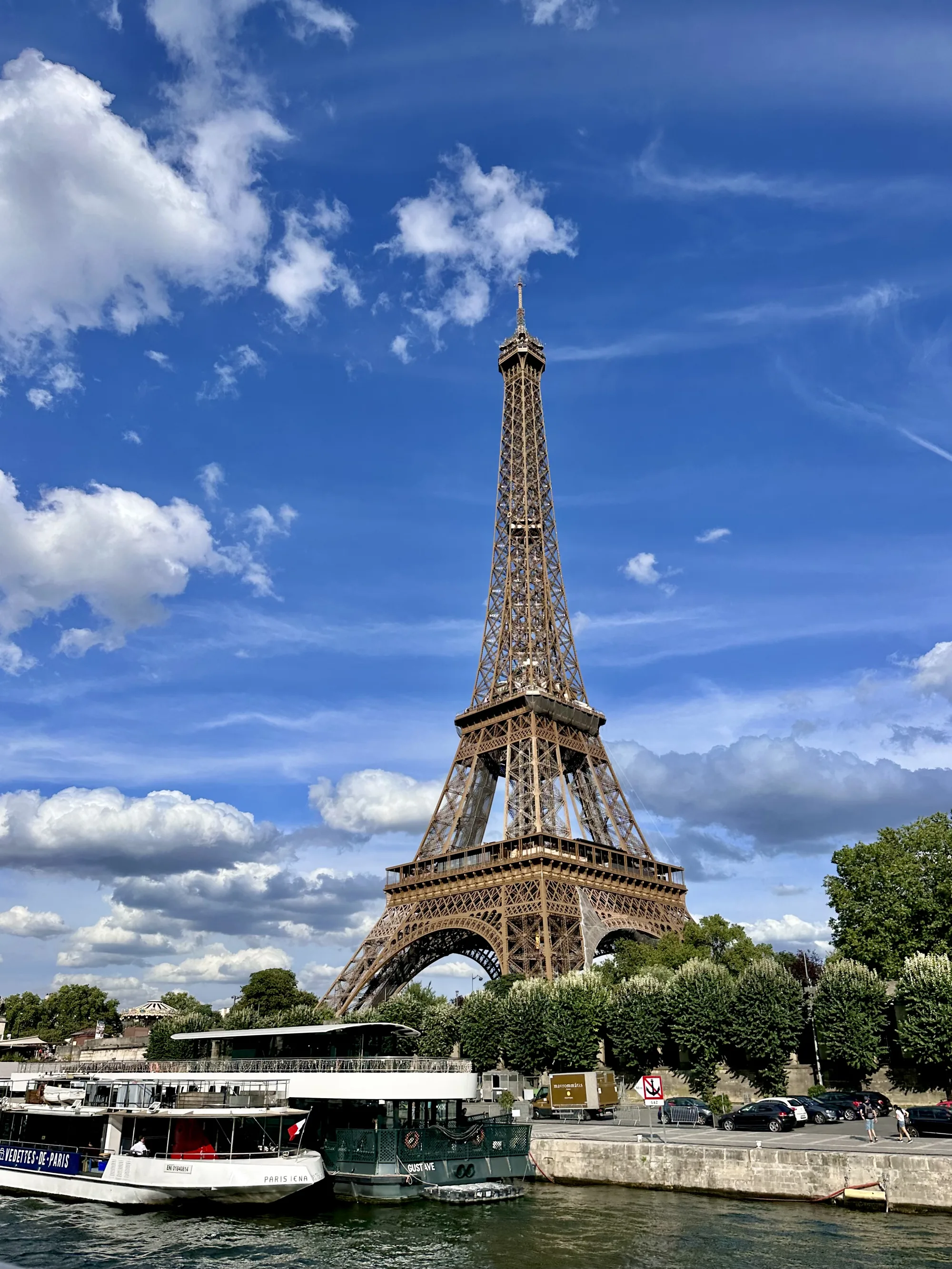 写真にはフランス・パリの象徴であるエッフェル塔が大きく写っています。青空を背景に、鉄骨で組まれたエッフェル塔が堂々とそびえ立っており、パリの美しい景観を象徴しています。空には白い雲が点在し、晴れた日の穏やかな雰囲気を醸し出しています。  エッフェル塔の足元にはセーヌ川が流れ、観光船が停泊しています。川岸には木々が茂り、緑豊かな風景が広がっています。川沿いの道路には車が停まっており、人々が歩いている様子も伺えます。全体的に、パリの美しい風景と観光客の賑わいが感じられる写真です。