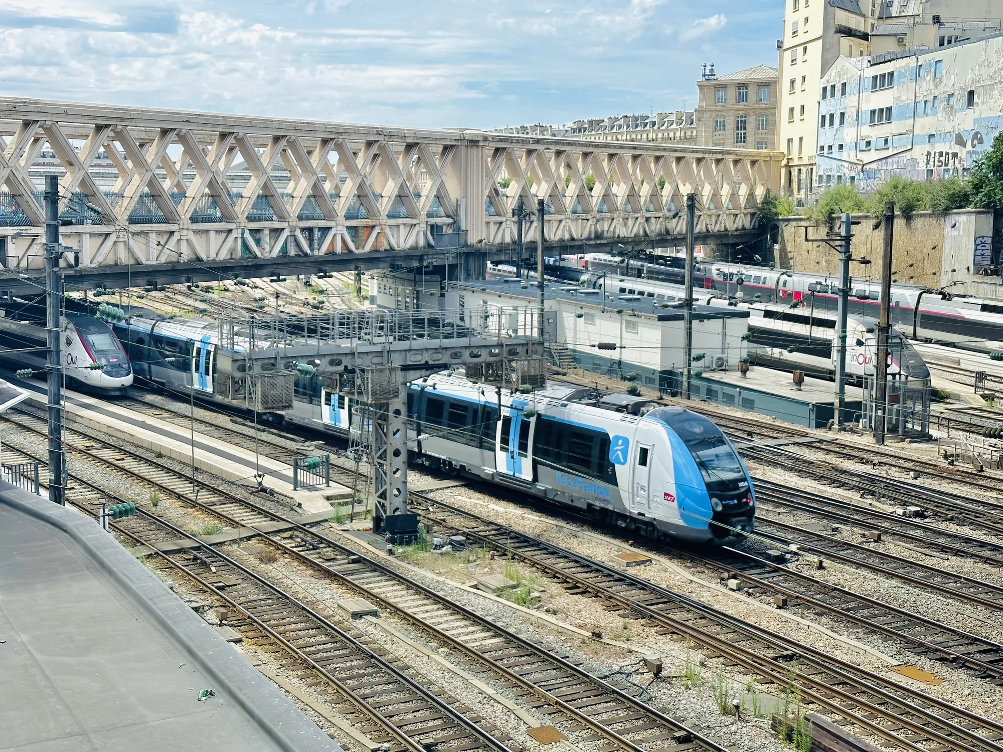 写真はフランスのパリにある鉄道駅の風景を捉えています。手前には無数の線路が敷かれており、その上をブルーとグレーのツートンカラーの近郊電車が走行しています。電車の車体には「Nord-France」と書かれています。奥には高速鉄道TGVの車両も見えます。上空には、特徴的な幾何学模様の鉄骨橋が架かっています。背景には、パリの街並みが見え、歴史的な建物が並んでいます。空は少し雲があり、明るい日中の光が全体を照らしています。鉄道施設特有の無機質さと、パリの街並みの調和が感じられる一枚です。