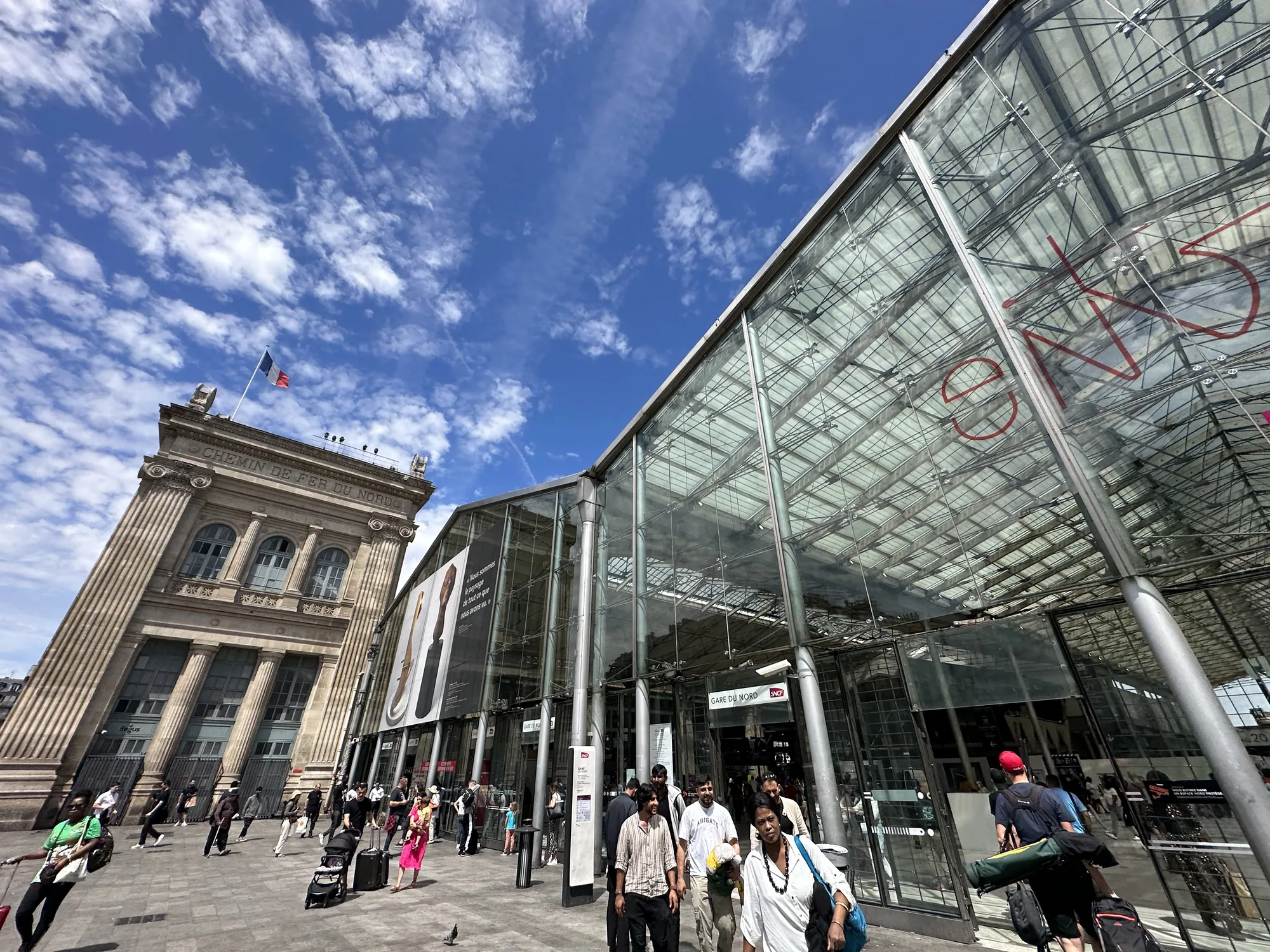 写真は、フランスのパリにあるパリ北駅（Gare du Nord）の駅舎正面をとらえたものです。青空が広がり、白い雲が浮かんでいます。画面左側には、フランス国旗が掲げられた歴史的な石造りの駅舎の一部が見えます。「CHEMIN DE FER DU NORD」の文字が刻まれています。画面右側には、近代的なガラス張りの駅舎が広がっています。ガラスには赤い文字で落書きのようなものが書かれています。駅舎の前には多くの人が行き交い、旅行者と思われる人々がスーツケースなどを持ち歩いています。駅の入り口には「GARE DU NORD」と書かれた看板があり、駅名が示されています。全体的に、活気があり、国際的な雰囲気を感じさせる写真です。