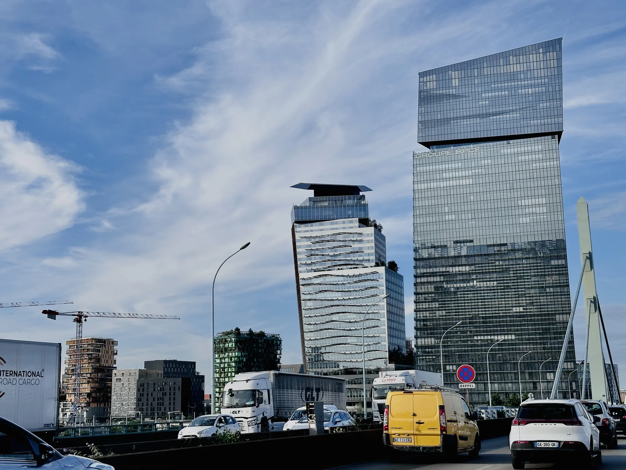 写真はフランスのパリで撮影されたもので、現代的な都市景観が写っています。空は青く、薄い雲が広がっています。道路には、トラック、乗用車、黄色いバンなど、様々な車両が走っています。背景には、ガラス張りの高層ビルがそびえ立ち、洗練された印象を与えています。高層ビルには「GROUPE BPE」の文字が見えます。建設中のクレーンも写っており、都市の発展を感じさせます。道路脇の標識やストリートライトも、都市の日常風景を捉えています。写真全体から、活気がありながらも落ち着いた雰囲気が伝わってきます。