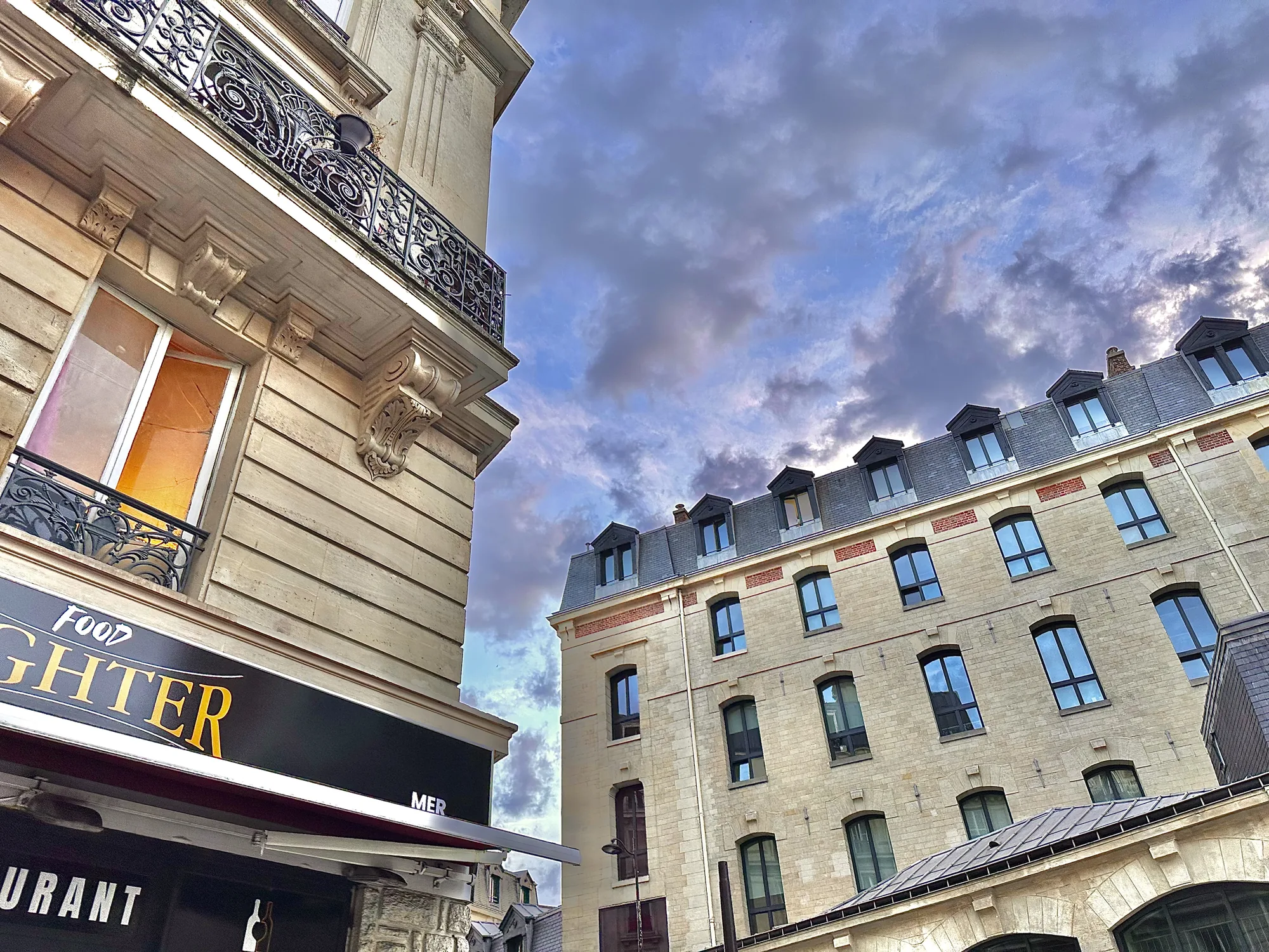 写真はフランスのパリで撮影されたもので、伝統的な石造りの建物が特徴的な街並みを捉えています。画面左側には、美しい装飾が施されたバルコニーを持つ建物が写っており、窓からは暖色の光が漏れています。建物には「FOOD FIGHTER RESTAURANT MER」と書かれた看板が見えます。  画面右側には、対照的に比較的シンプルな外観の建物がそびえ立っており、複数の窓が並んでいます。屋根にはドーマー窓が配置され、独特の雰囲気を醸し出しています。  空にはドラマチックな雲が広がり、建物の間から見える空模様が印象的です。全体的に、パリの歴史的な美しさと現代的な生活が調和している様子が伝わってくる写真です。