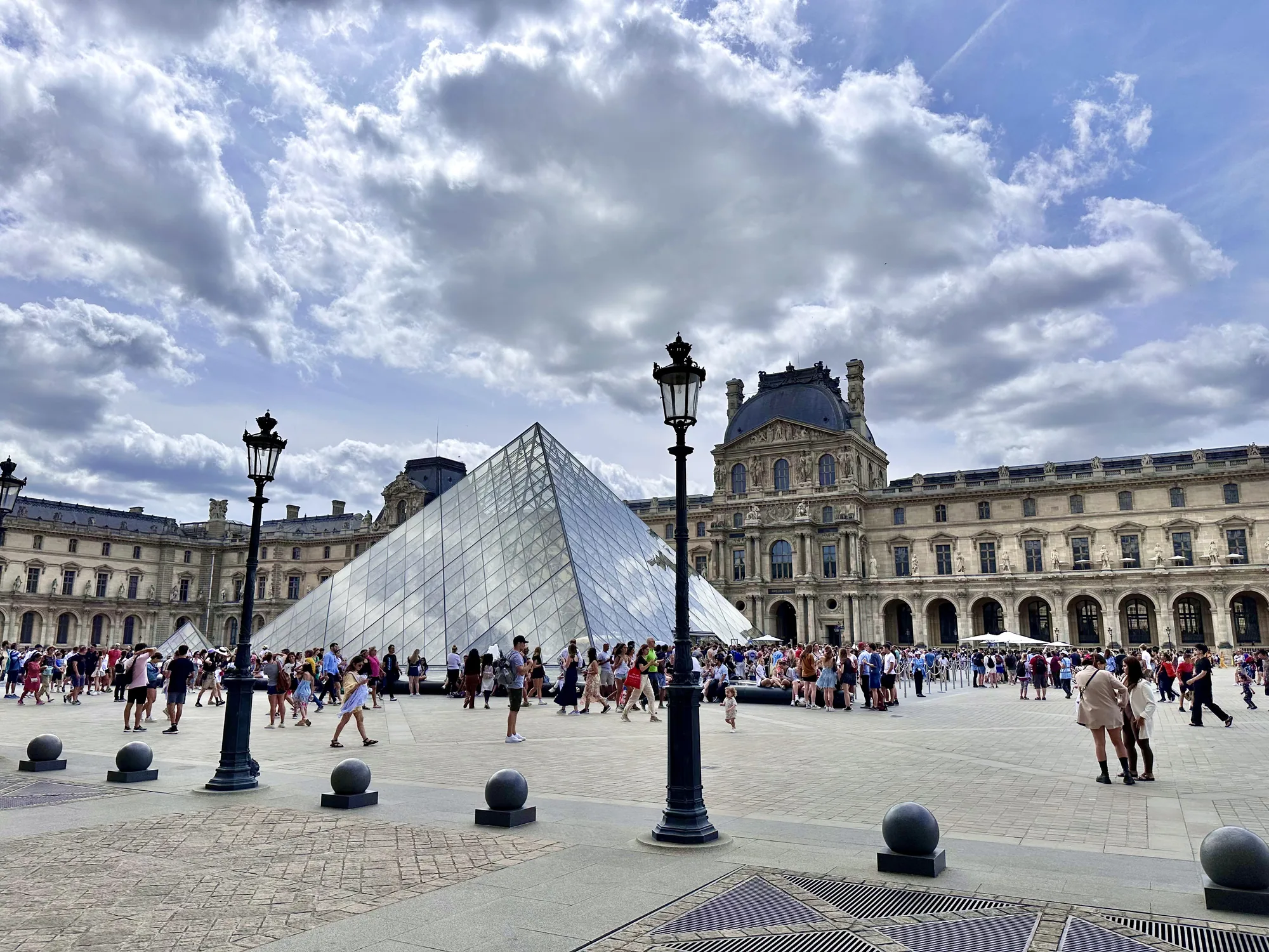この写真はフランスのパリにあるルーブル美術館の前で撮影されたものです。写真には、ガラス製のピラミッド型のモニュメントが大きく写っており、ルーブル美術館の象徴的な存在感を示しています。ピラミッドの前には広場が広がり、多くの観光客が行き交っています。人々は様々な服装をしており、その表情は様々です。広場には、丸い石のオブジェや歴史を感じさせる街灯が配置されており、美しい景観を形成しています。背景には、ルーブル美術館の壮大な建物がそびえ立ち、その歴史的な重みを感じさせます。空には、厚い雲が広がっており、独特の雰囲気を作り出しています。全体として、活気に満ちた観光地の様子と、歴史的な建造物の荘厳さが組み合わさった、印象的な一枚です。