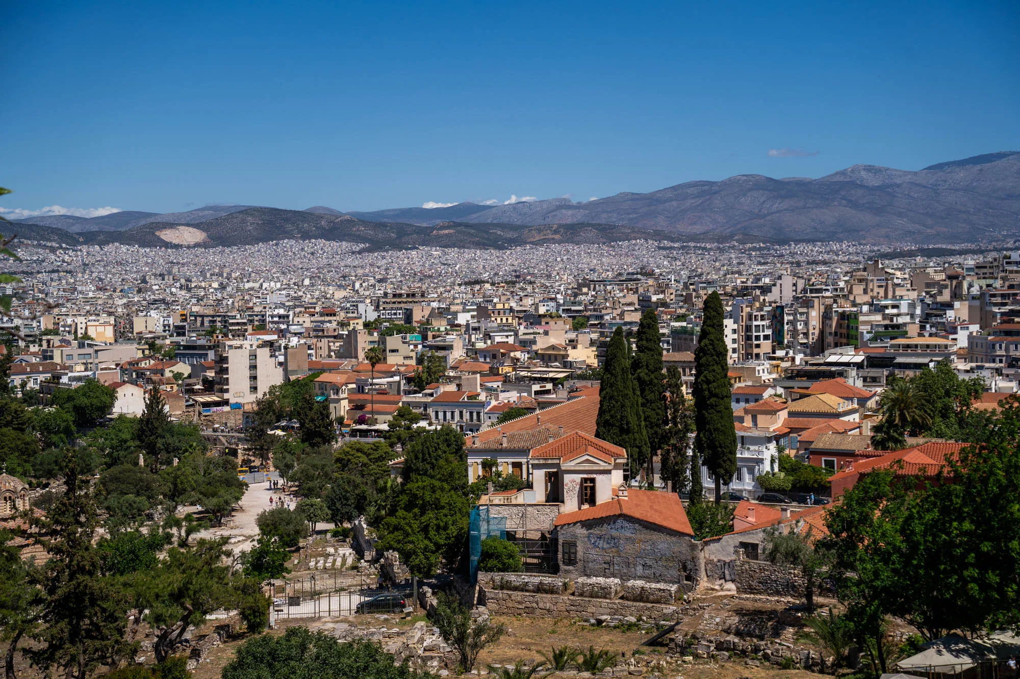 ギリシャのアテネで撮影された、街並みを見下ろす風景写真です。手前には緑豊かな木々や、赤茶色の屋根の建物がいくつか見えます。それらの建物は比較的小規模で、歴史を感じさせる風合いです。その奥には、高層ビルやアパートが密集したアテネの市街地が広がっています。建物が隙間なく立ち並び、都市の規模の大きさを感じさせます。さらに遠くには、青みがかった山々が連なっており、空の青さと相まって、美しい背景を形成しています。空は晴れ渡っており、明るい日差しが街全体を照らしています。全体的に、歴史的な建造物と現代的な都市が融合した、アテネの独特な風景を捉えた写真です。