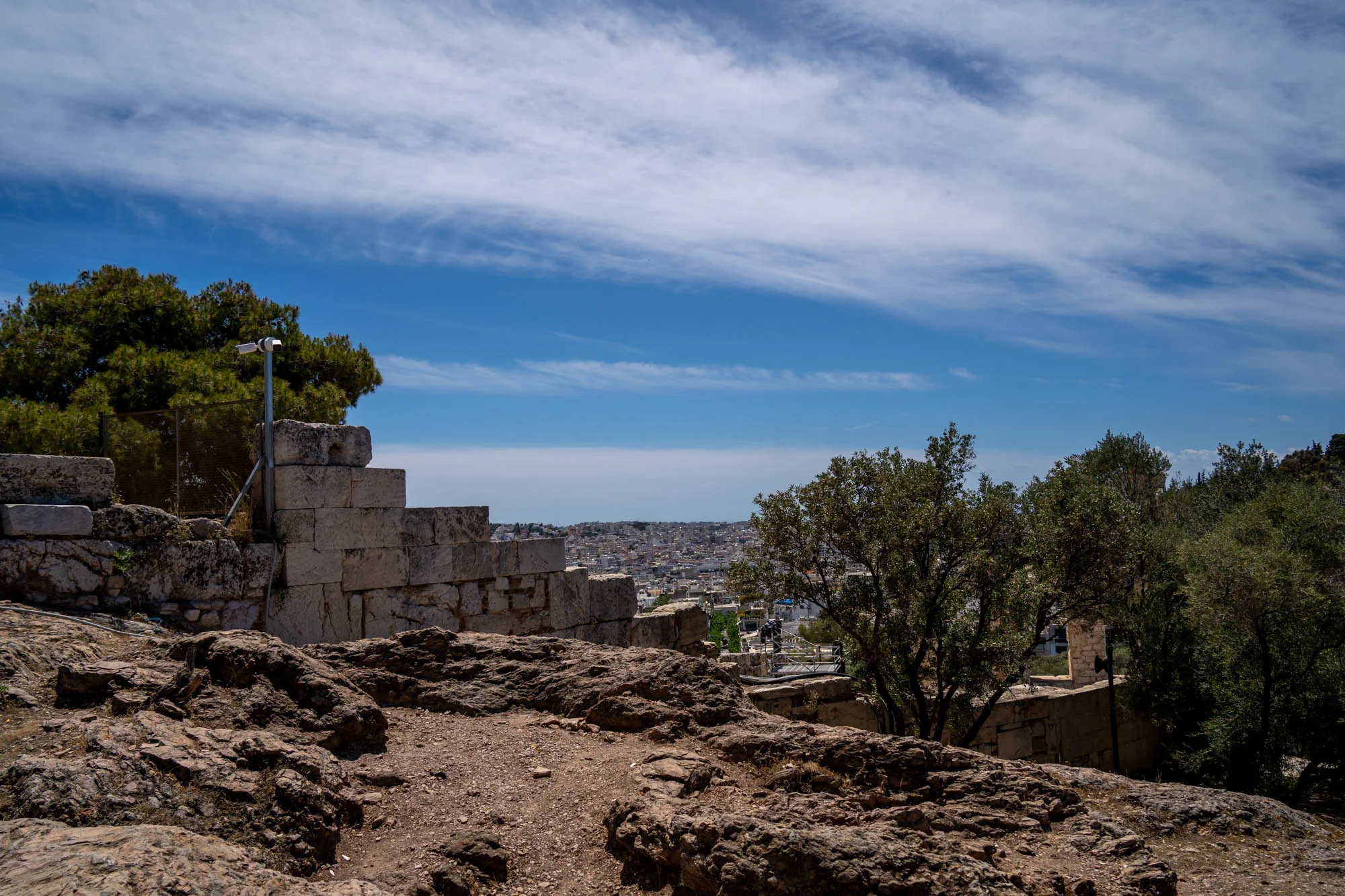 ギリシャのアテネで撮影された写真です。前景には、岩がゴツゴツとした地面が広がっています。その奥には、積み上げられた石垣の遺跡が見えます。石垣のそばには緑の木々が生い茂り、その間には金属製のフェンスと監視カメラが設置されています。遠景にはアテネの街並みが広がり、建物が密集している様子が伺えます。空は青く、白い筋状の雲が流れています。全体的に、古代遺跡と現代の都市が共存する風景が捉えられており、歴史と自然、そして現代的な要素が組み合わさった独特の雰囲気を醸し出しています。