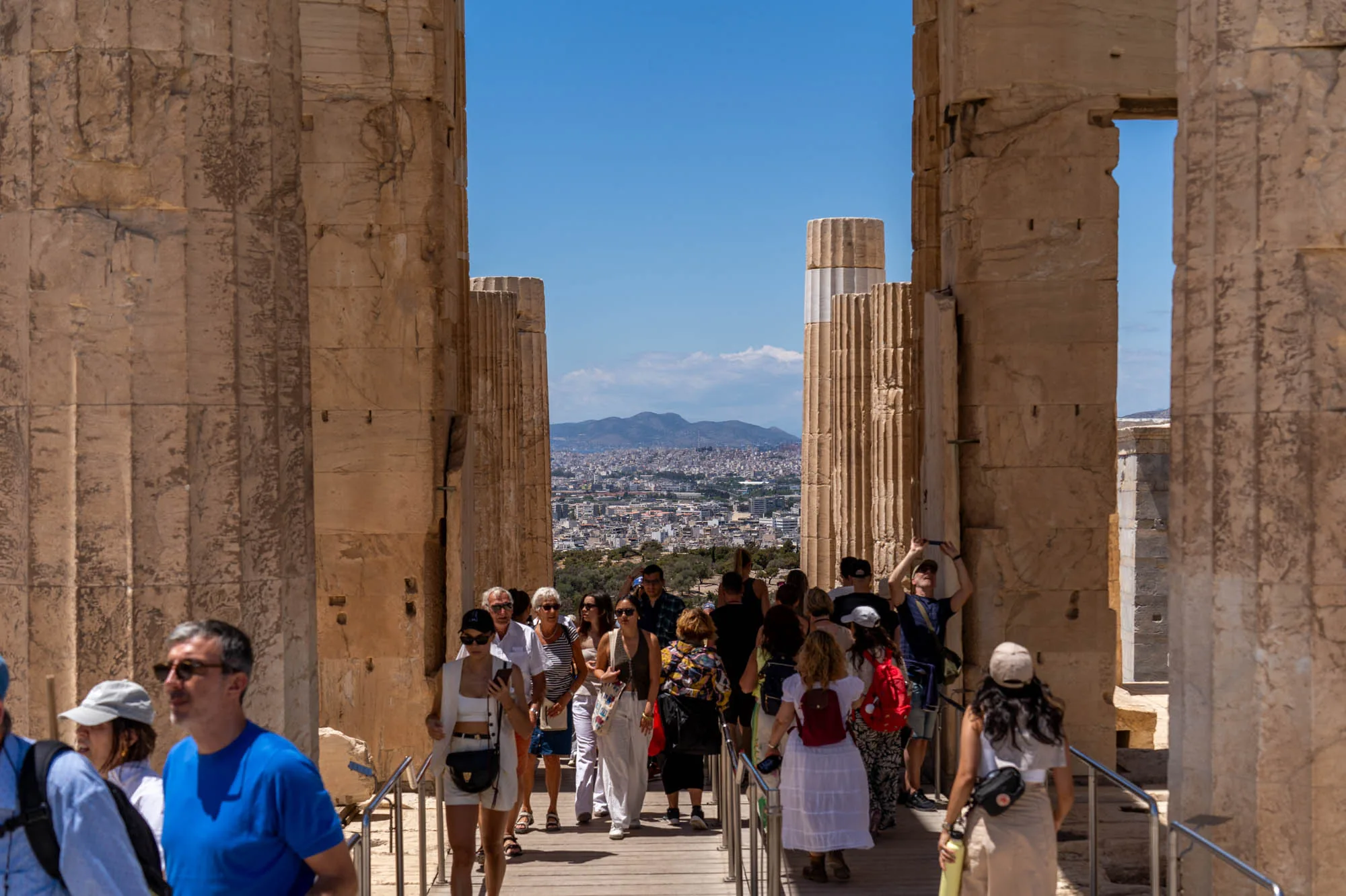 ギリシャのアテネで撮影された写真です。古代遺跡の一部である石柱が複数立っており、その間を観光客が大勢歩いています。柱の隙間からはアテネの市街地が見え、遠くには山々が連なっています。空は晴れ渡っており、日差しが強く、観光客はそれぞれ日焼け対策をしているようです。遺跡の歴史的な雰囲気と、観光客の活気が混ざり合った写真です。