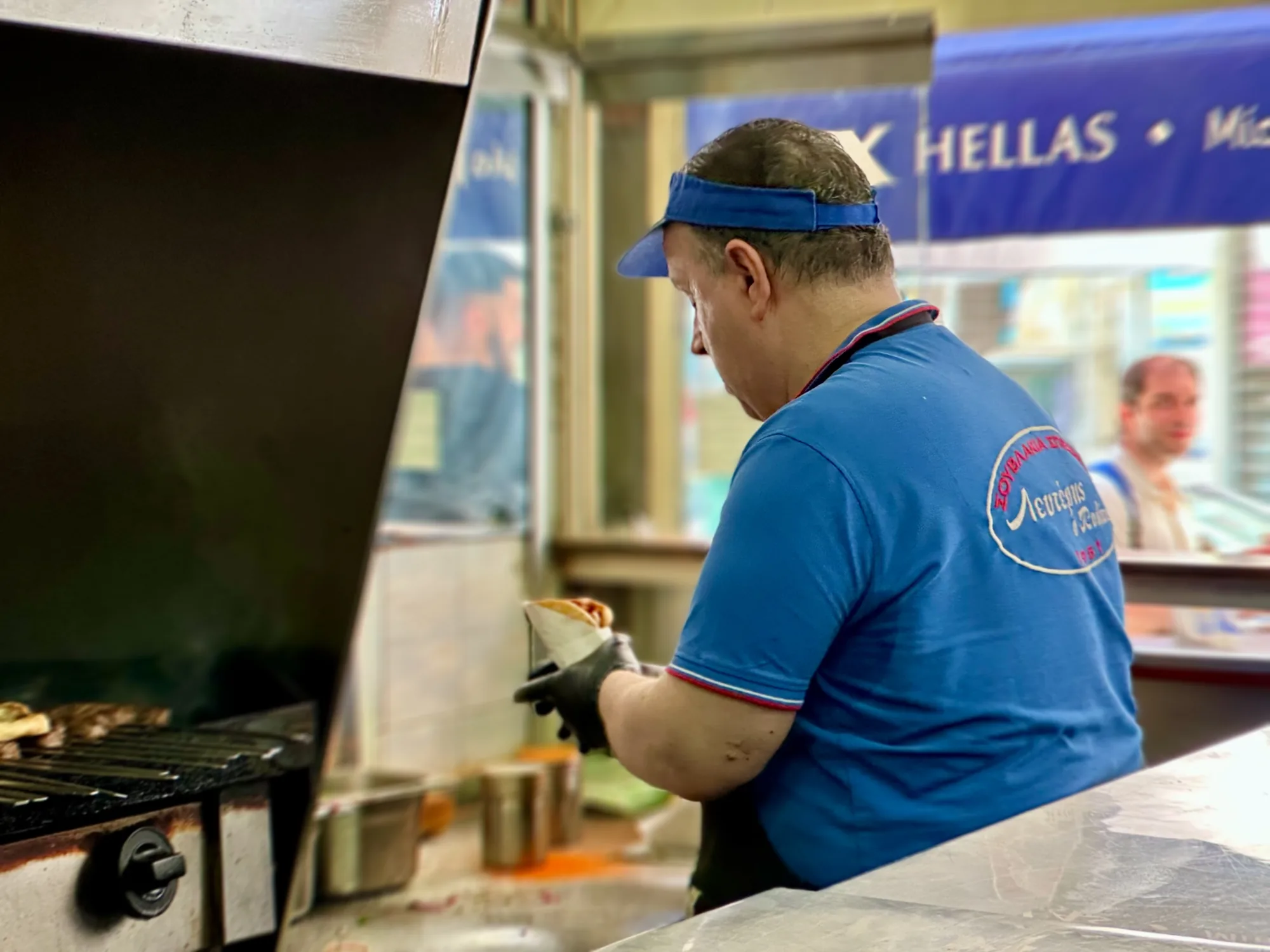 この写真はギリシャのアテネで撮影されたと思われる屋台の様子を捉えています。手前に見えるのは調理台とグリルで、ソーセージのようなものが焼かれています。調理人と思われる男性が黒い手袋を着用し、何かの料理を紙で包んでいます。青いシャツを着ており、背中には白い円形のロゴのようなものがあり、ギリシャ語で「Λευτέρης Ο Κωστα」のような文字が見えます。男性は青いバイザーキャップを被っています。背景には別の男性の姿がぼんやりと見え、屋台の看板には「HELLAS」という文字が確認できます。全体的に活気のある屋台の雰囲気が伝わってくる一枚です。