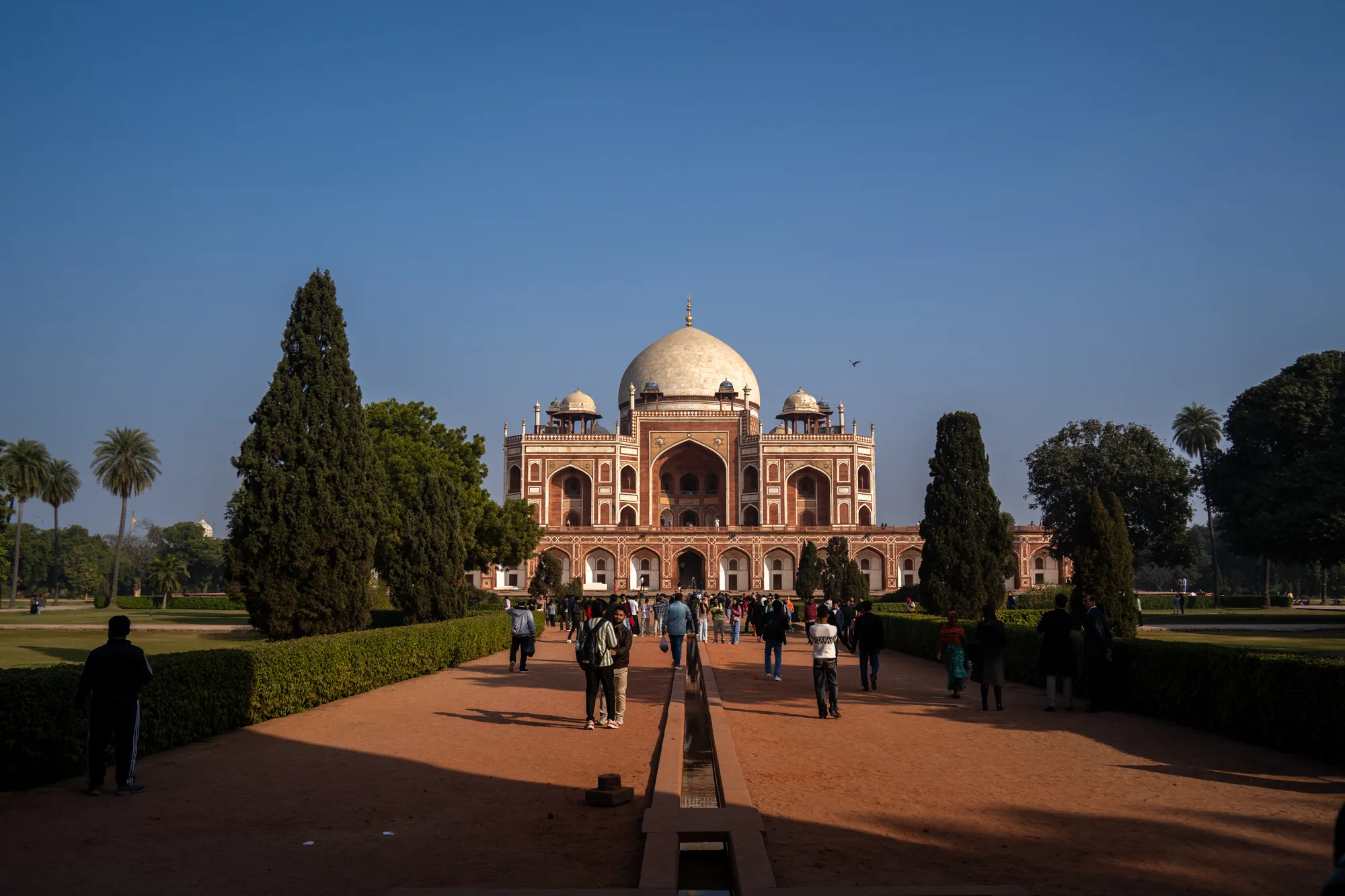 インド、ニューデリーにあるフマユーン廟を捉えた写真です。明るい青空の下、堂々とした廟が中央に位置しています。廟は赤茶色と白のコントラストが美しい、精巧な装飾が施された建物で、中央には大きな白いドームがあります。廟の前には、手入れの行き届いた庭園が広がっており、低い生垣や、まっすぐ伸びる水路が配置されています。庭園を歩く多くの人々が写っており、観光地としての賑わいを感じさせます。写真全体の色調は暖かく、歴史的な建造物の重厚さと、人々の活気が調和した印象を与えます。画面の左右には緑豊かな木々が配置され、奥行きを演出しています。