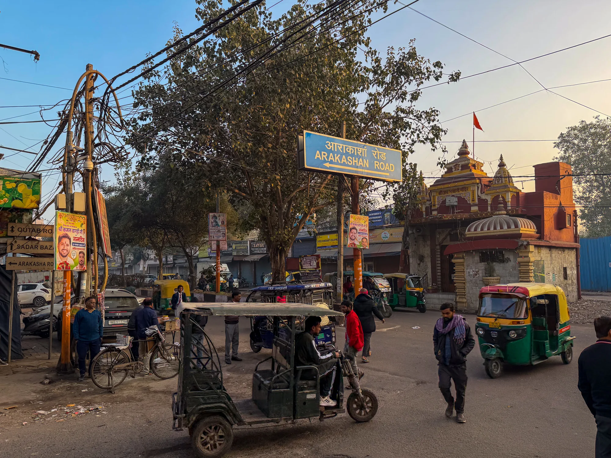 インドのニューデリーで撮影された写真です。街の一角の様子が写っており、道路標識には「ARAKASHAN ROAD」と書かれています。道路にはオートリクシャー（タクシー）が複数台走っており、人々が歩いています。電線が複雑に張り巡らされており、インドらしい混沌とした風景が印象的です。道端には自転車や広告、露店のようなものも見られます。背景には寺院のような建物が確認できます。全体的にやや埃っぽく、生活感あふれる街の雰囲気が伝わってきます。