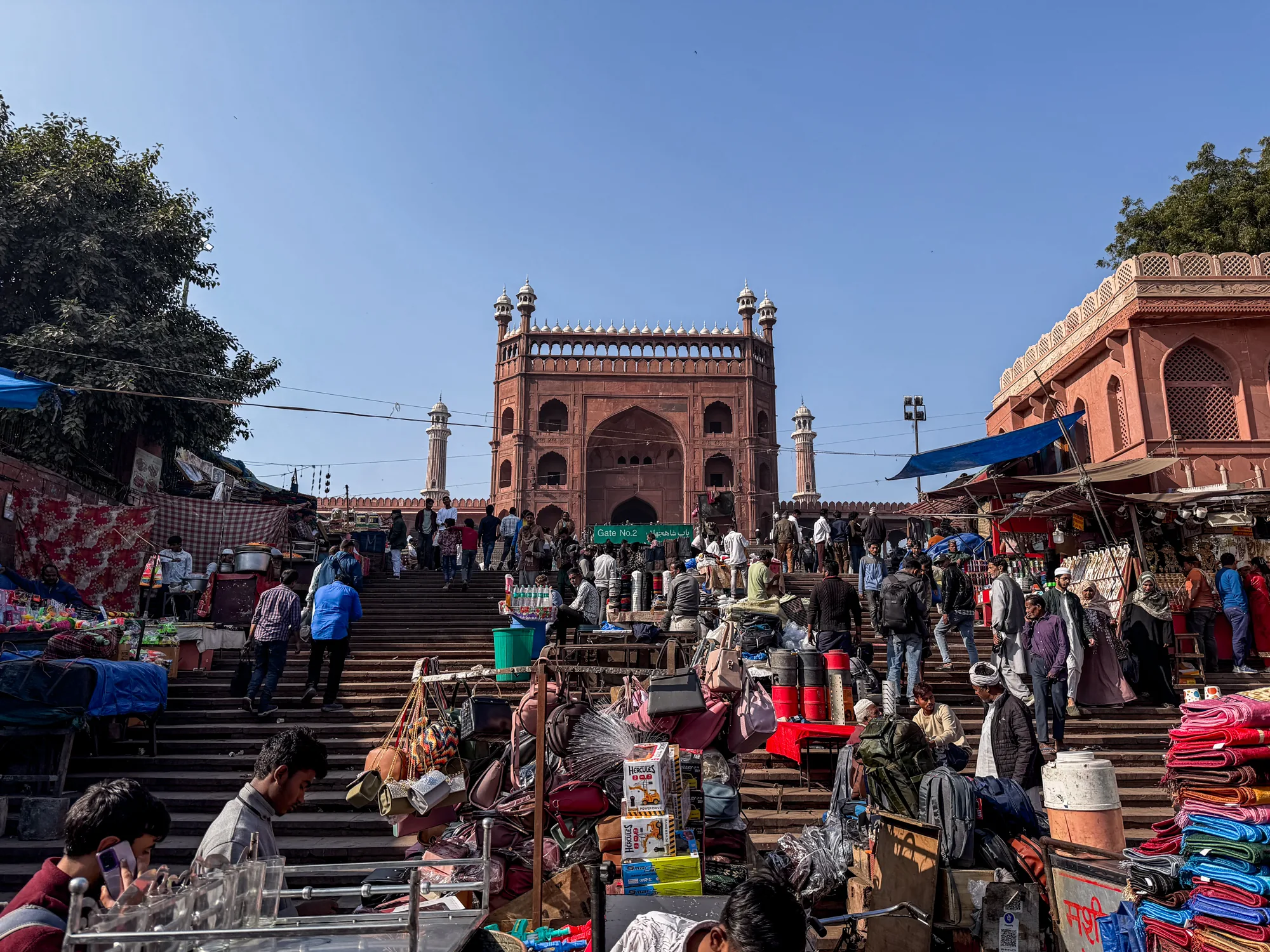 写真はインドのニューデリーで撮影されたもので、賑やかな光景が広がっています。中央には赤茶色の砂岩でできた壮大なモスクの門が見え、その前に広がる階段は多くの人々で埋め尽くされています。階段の両側には露店が立ち並び、バッグ、布地、おもちゃなど様々な商品が所狭しと並べられています。人々は階段を上り下りしたり、露店をのぞいたりしており、活気に満ちた雰囲気が伝わってきます。空は晴れており、建物や人々のシルエットがくっきりと浮かび上がっています。露店の屋根には青いシートがかかっており、日差しを遮っています。モスクの門の左右には、同じ赤茶色の建物が立ち、全体の景観に統一感を与えています。人々の服装も様々で、伝統的な衣装を着た人もいれば、現代的な服装の人もいます。全体として、デリーの活気あふれる日常を切り取った一枚と言えるでしょう。