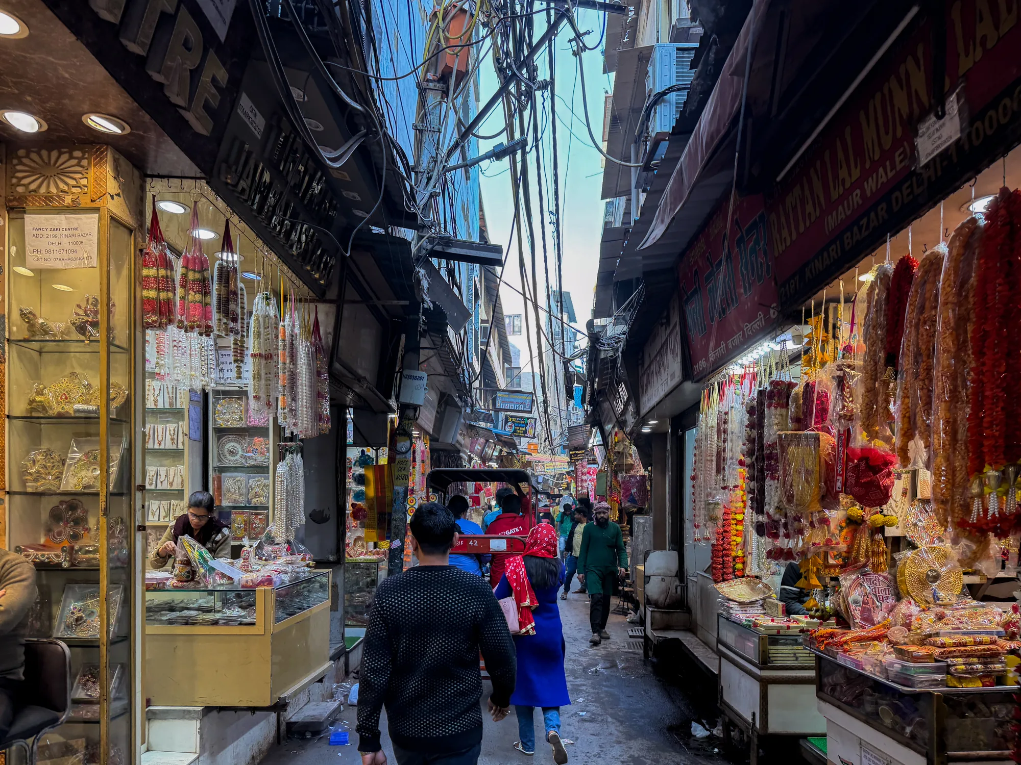 この写真は、インドのニューデリーにある市場の様子を捉えたものです。狭い路地の両側には店が軒を連ね、それぞれの商品を所狭しと並べています。左側の店には、装飾品やアクセサリーがガラスケースに展示され、店員が手入れをしている様子が見えます。右側の店は、花輪や装飾品が店の軒先に吊り下げられ、カラフルな商品が目を引きます。路地は人で賑わい、歩行者やリクシャーが行き交っています。頭上には電線が複雑に絡み合い、空を覆い隠すように張り巡らされています。建物の壁は古びており、市場の歴史を感じさせます。全体的に活気があり、エネルギーに満ちた雰囲気の写真です。