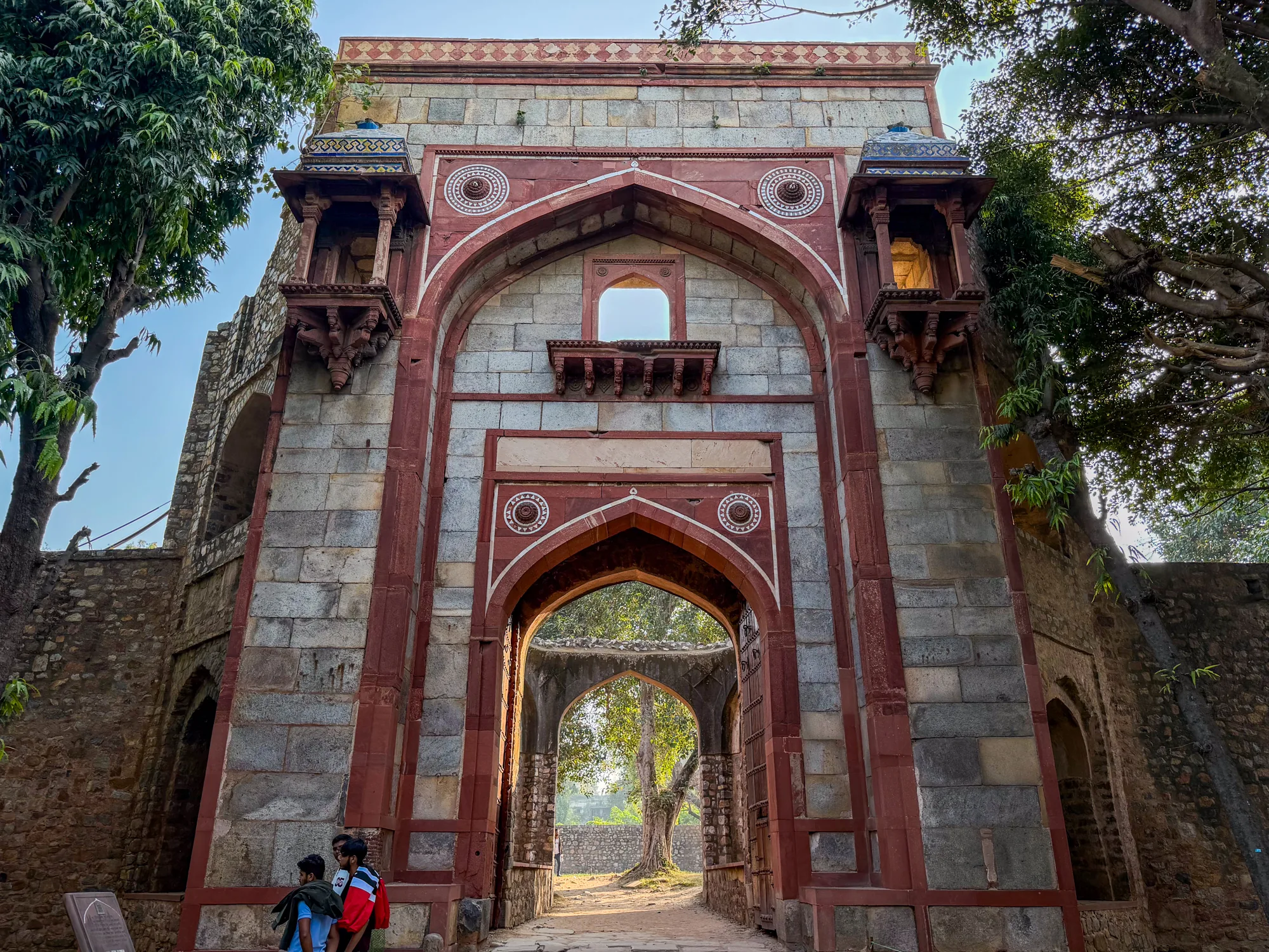 写真は、インドのニューデリーで撮影されたと思われる、歴史的な建造物の門を写しています。門は赤とグレーの石で作られており、アーチ型の入り口が特徴的です。門の上部には小さなバルコニーのような装飾が施され、細部まで丁寧に作られている様子が伺えます。門の内側には、さらに奥へと続くアーチ型の構造が見え、遠くには木々が見えます。門の前には数人の人物が立っており、建物の大きさを際立たせています。全体的に、歴史的な重みと静けさを感じさせる写真です。日差しはやや強く、門の影を濃くしています。周りの木々がさらに歴史的な雰囲気を醸し出しています。