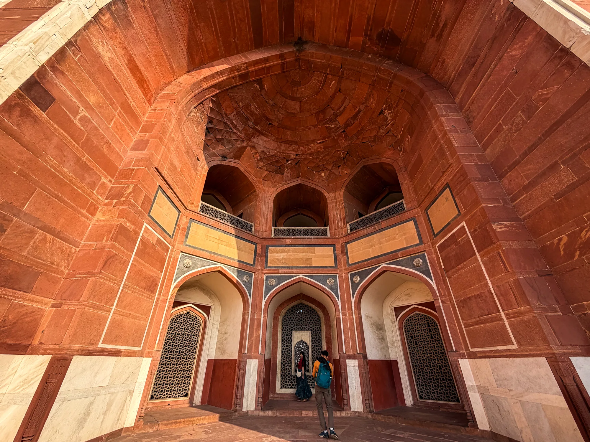 インド、ニューデリーで撮影された写真です。カメラはローアングルから、壮大な建築物の内部を捉えています。建物は赤レンガ造りで、力強いアーチ構造が印象的です。入り口は複数あり、幾何学的な模様が施された石の格子窓がはめ込まれています。中央の入り口には2人の人物が立っており、一人は赤いトップスに柄物のスカートを着用し、もう一人は黄色いシャツにリュックサックを背負っています。地面には光が差し込み、赤レンガの色をより鮮やかにしています。全体的に、歴史的な重厚感と静けさが感じられる写真です。