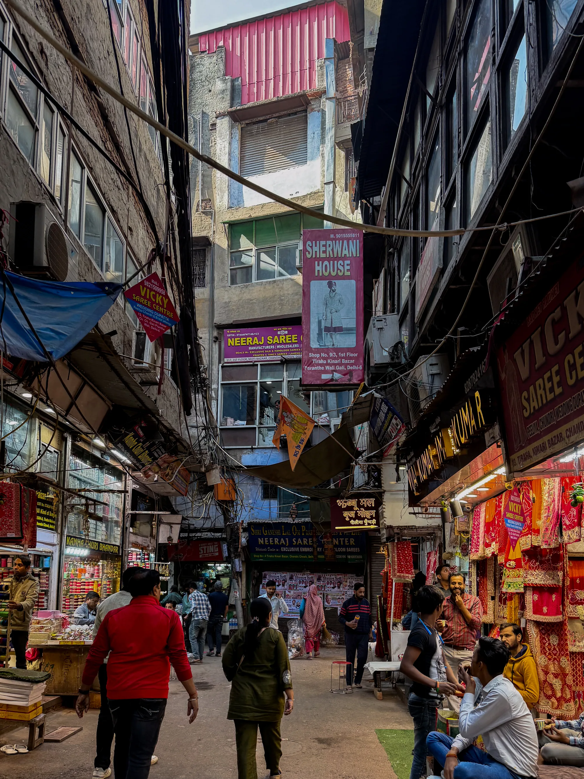 この写真はインドのニューデリーにある市場の賑やかな通りを捉えています。狭い路地には人々が行き交い、両側には所狭しと店舗が並んでいます。店舗にはサリーや伝統的な衣装が展示され、鮮やかな色彩が目を引きます。看板や広告がひしめき合い、電気ケーブルが空を覆い、混沌とした雰囲気を醸し出しています。人々は歩いたり、立ち止まって話をしたり、買い物をしたりと様々です。路地は薄暗く、店舗からの明かりが商品を照らし出しています。写真全体から、活気と雑多な魅力が感じられます。