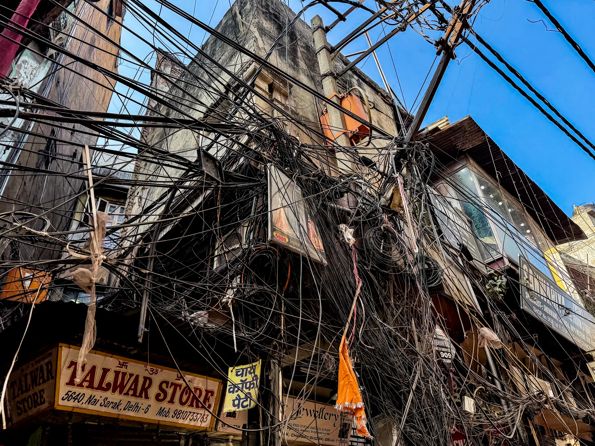 インドのニューデリーで撮影された写真です。画面全体に、建物の外壁を覆い尽くすように無数の電線が張り巡らされています。電線は黒く、複雑に絡み合い、空の青さをわずかに覗かせています。建物の壁は古びており、汚れやひび割れが見られます。建物の下には店舗があり、「TALWAR STORE」や「Jewellery」などの看板が見えます。オレンジ色の旗のようなものもぶら下がっています。電線の錯綜ぶりと、古びた建物の様子が、ニューデリーの混沌とした雰囲気を伝えています。