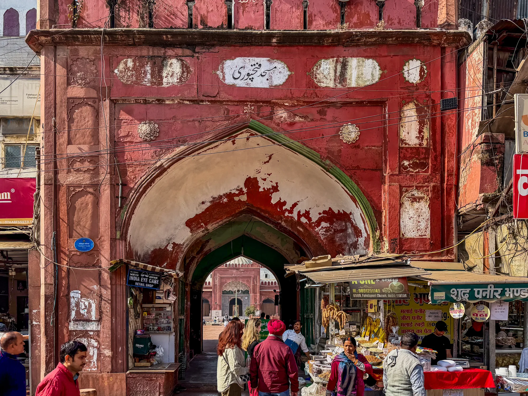 インド、ニューデリーで撮影された写真です。焦点となっているのは、赤茶色の石造りの大きなアーチ型の門です。門の上部には装飾的な模様とアラビア文字のような碑文が見られます。壁の表面は風化しており、歴史を感じさせる趣があります。アーチの奥には、同様の赤茶色の建物が見え、おそらくモスクなどの宗教施設でしょう。門の左右には、露店が立ち並び、賑やかな様子が伝わってきます。露店には様々な商品が並べられており、人々が買い物を楽しんでいるようです。写真全体として、インドの歴史と文化、そして人々の生活が混ざり合った風景を描き出しています。太陽の光が差し込み、活気ある雰囲気を醸し出しています。手前には人々が歩いており、そのうち何人かはカメラの方を見ています。