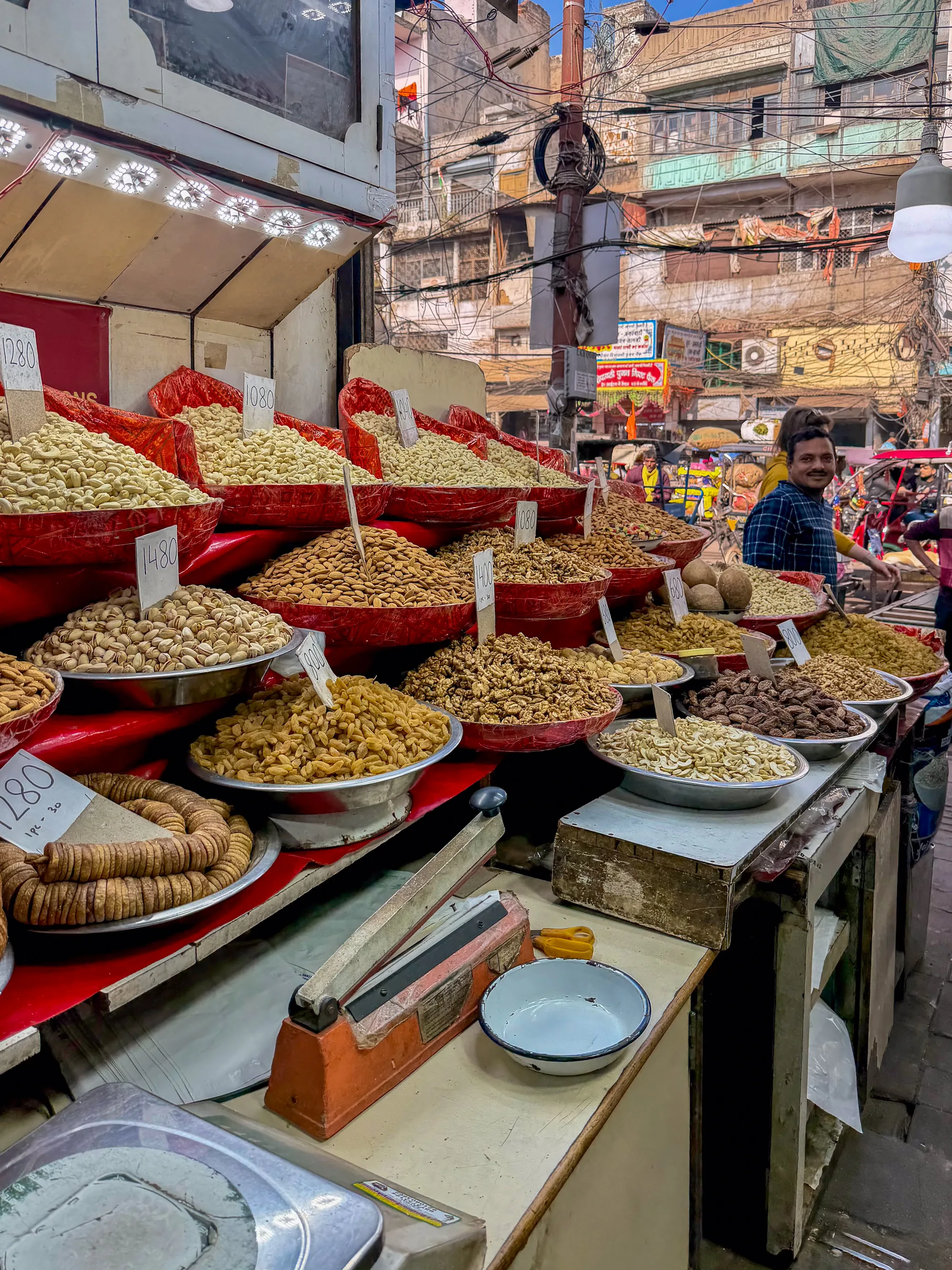 写真はインドのニューデリーで撮影された、ナッツやドライフルーツを売る店先の様子を写しています。店先には大量のナッツが盛られており、アーモンド、カシューナッツ、クルミ、レーズン、デーツ、イチジクなどが、赤い器やステンレスのボウルに山積みにされています。それぞれの容器には手書きの価格が表示された小さな札が立てられています。  店員の男性が笑顔でこちらを見ており、背景には他の買い物客やお店、そして複雑に絡み合った電線が見えます。電線はインドの街並みによく見られる光景で、建物の外壁を這うように張り巡らされています。店の照明は暖色系のライトで、商品に照りを与え、食欲をそそるように演出しています。店先は活気にあふれ、インドの市場の賑やかさを伝えています。全体的に活気ある市場の雰囲気が伝わる一枚です。