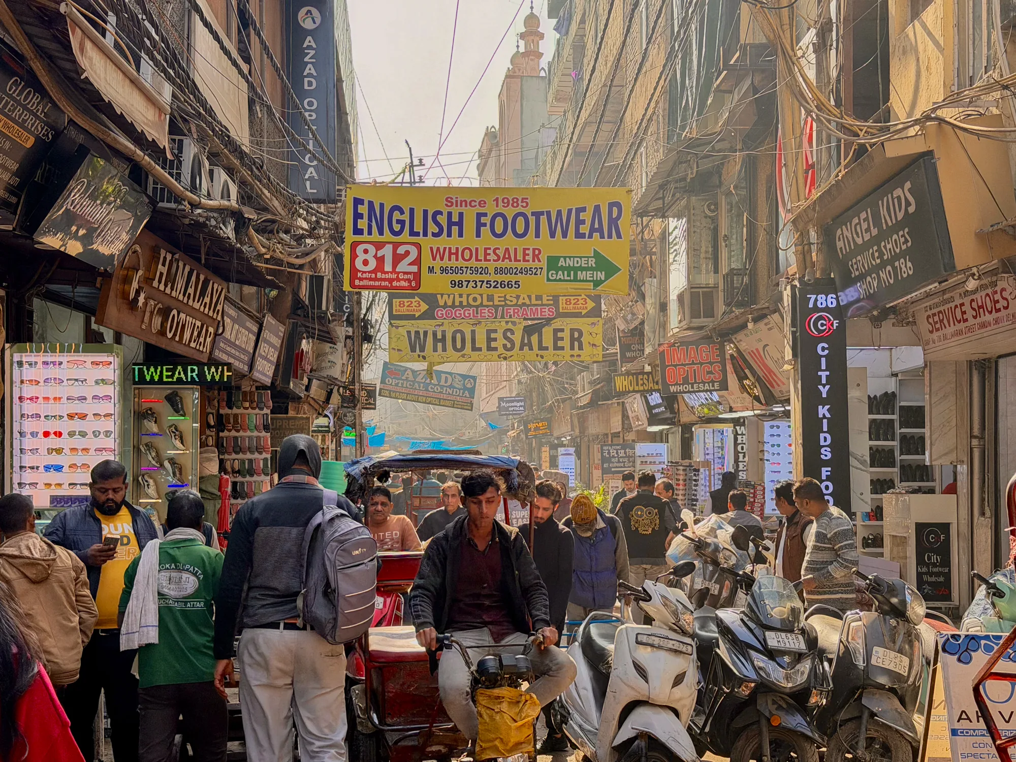 写真はインドのニューデリーで撮影された、賑やかな通りを写したものです。道は人で賑わい、リキシャ、オートバイなどの交通手段が混在しています。通り沿いには、様々な商品を販売する商店が軒を連ねています。看板には「ENGLISH FOOTWEAR」「ANGEL KIDS」「HIMALAYA FOOTWEAR」などと英語で書かれたものが見られます。電線が複雑に絡み合い、建物の上空を覆っているのが印象的です。写真全体から、活気と混沌が入り混じった、インドらしいエネルギーを感じ取れます。街並みは歴史を感じさせる古い建物と、現代的な商業が混在しており、独特の雰囲気を醸し出しています。人々の服装も様々で、伝統的な衣装を着た人もいれば、現代的な服装の人もいます。全体的に暖色系の色調で、埃っぽく、活気のあるインドの日常を切り取った一枚です。