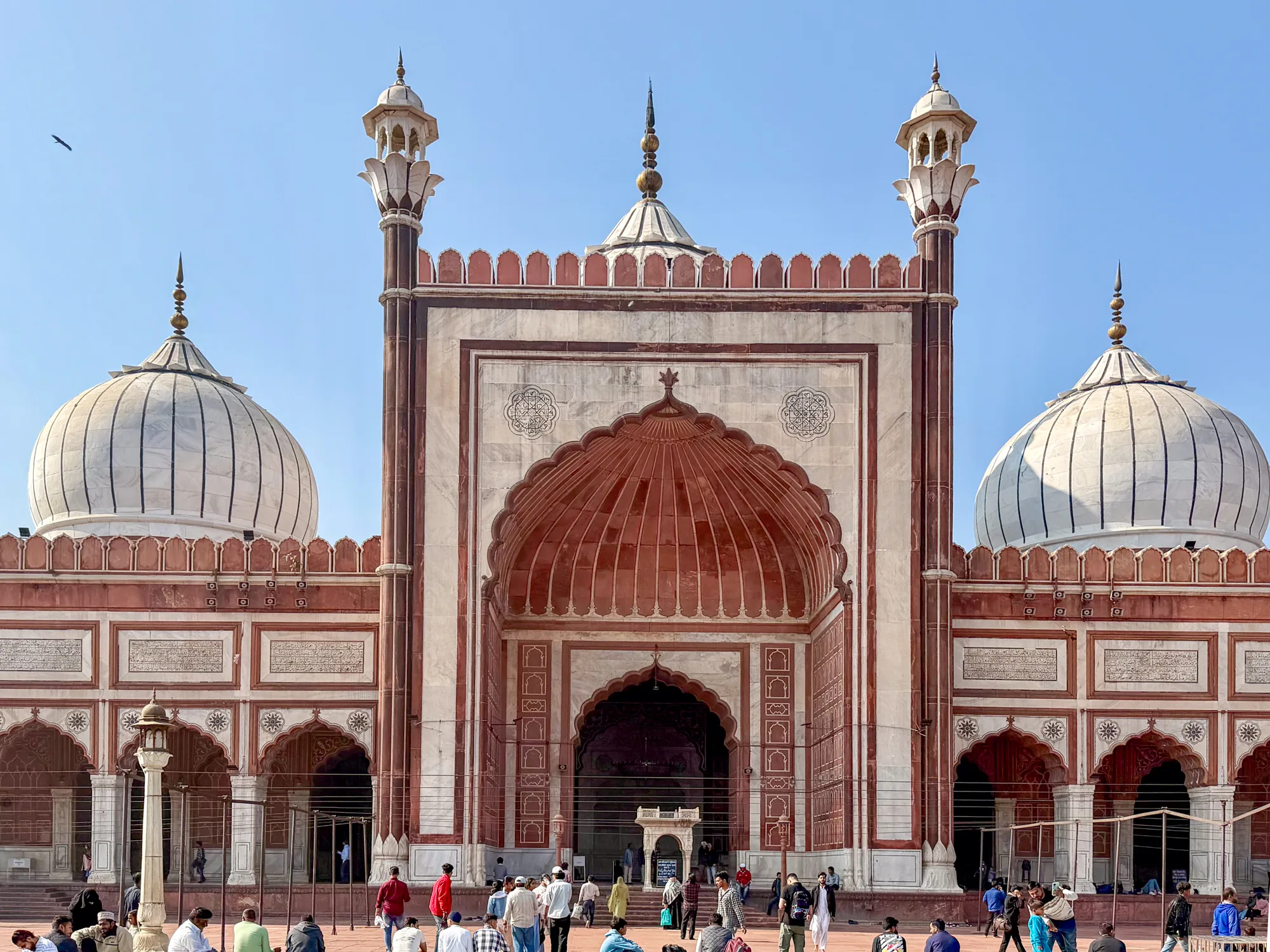写真はインドのニューデリーにある、壮大なモスクの正面を捉えています。空は晴れ渡っており、建物の赤茶色と白の配色が際立っています。モスクの入口は巨大なアーチ型で、細部にまでこだわった装飾が施されています。両脇にはドーム型の屋根がそびえ立ち、頂点には尖塔が立っています。建物全体は、幾何学的な模様やアラビア文字の装飾で飾られています。  モスクの前には、人々が集まっており、祈りを捧げたり、談笑したり、休憩したりしている様子が見られます。彼らの服装は様々で、伝統的なイスラムの衣装を着ている人もいれば、カジュアルな服装の人もいます。地面は石畳で舗装されており、遠くまで広がっています。全体的に、写真からは、宗教的な雰囲気と人々の活気が感じられます。鳥が空を舞い、遠くまで続く空の青さが、より一層、被写体のモスクの美しさを際立たせています。