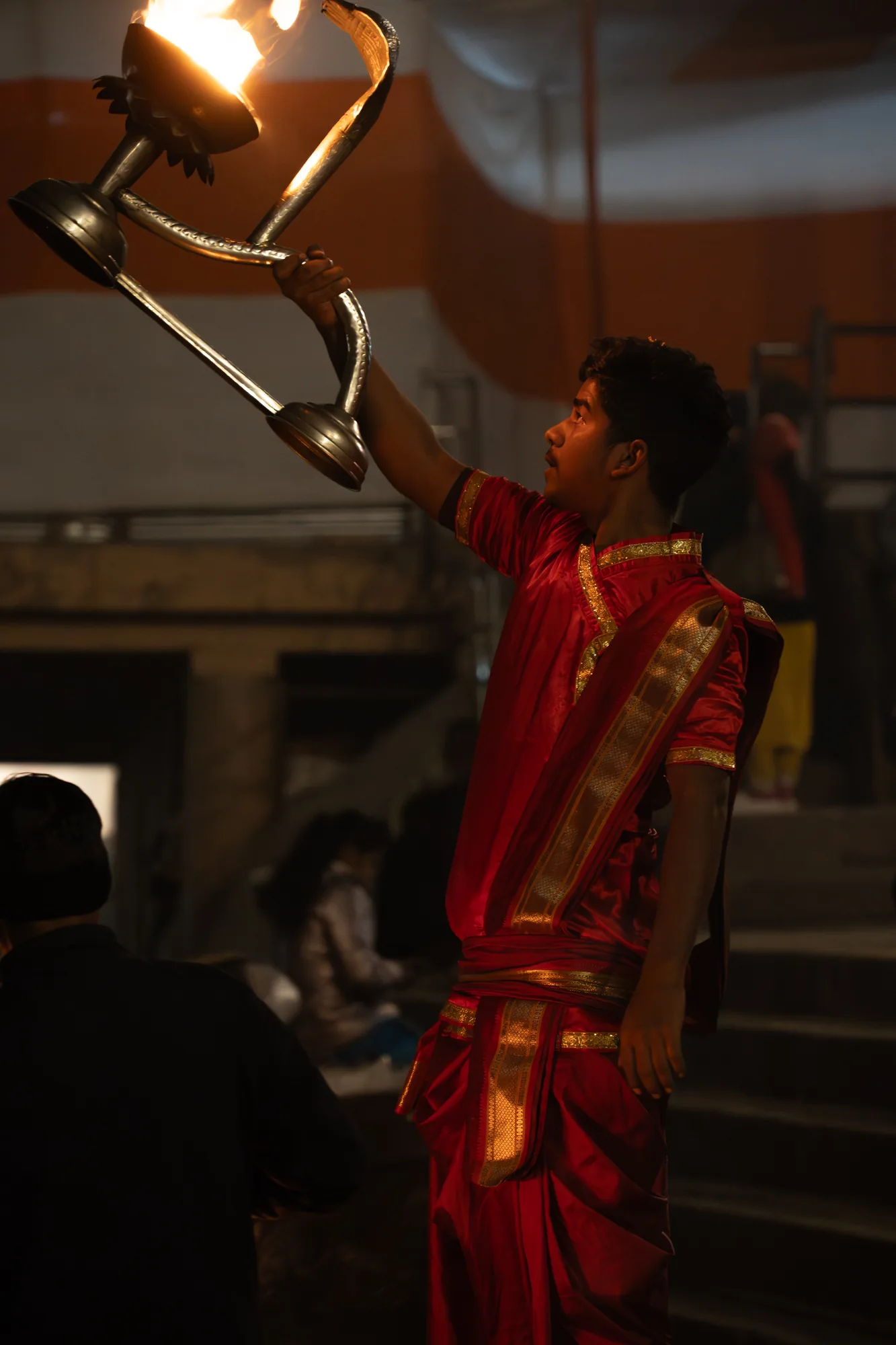 インドのバラナシで撮影された写真です。暗い背景の中で、赤い衣装を身につけた若い男性が、燃え盛る炎を捧げる儀式を行っています。男性は伝統的な赤い衣装を身につけ、金色の装飾が施されています。彼は右腕を高く上げ、複雑な金属製の器具を持ち、その上には燃える炎が立ち上っています。彼の表情は真剣で、儀式に集中している様子が伺えます。背景には他の人々がいますが、焦点は男性とその捧げ物に当てられています。全体的に、この写真はインドの宗教的儀式の神聖さと厳粛さを捉えています。