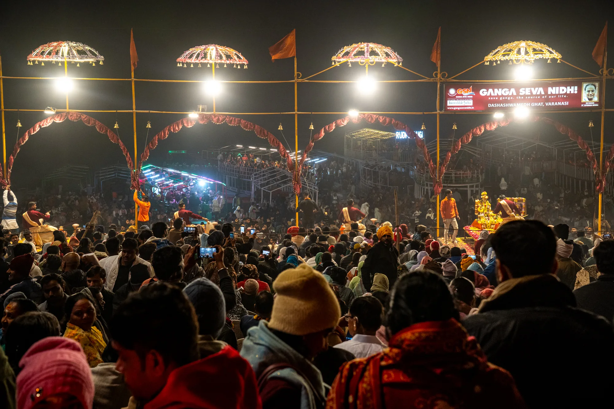 インド、バラナシのダシャーシュワメード・ガートで撮影された夜の写真です。群衆が密集し、人々が祭りの様子を見守っています。多くの人がスマートフォンで写真やビデオを撮影しています。前景には人々の頭部が多数見られ、様々な服装をしています。背景には、装飾されたアーチや照明が設置されており、祭りの雰囲気を高めています。「GANGA SEVA NIDHI!」と書かれた看板が見えます。これは、ガンジス川の保全を訴える団体の名前のようです。全体的に、活気と熱気に満ちた雰囲気で、宗教的な祭りの様子が伝わってきます。