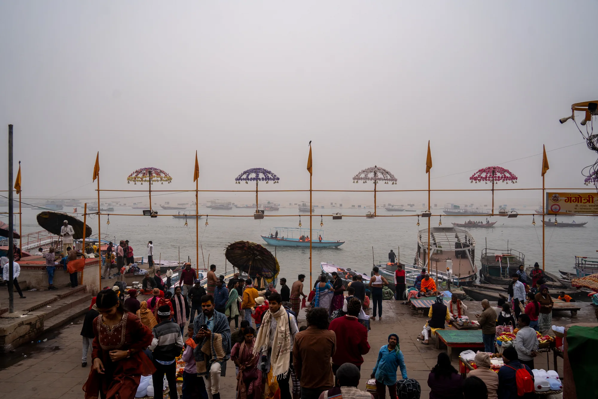 この写真は、インドのバラナシで撮影された、ガンジス川のほとりの賑わいを捉えた一枚です。 画面全体は、霧がかったような、あるいは霞がかかったようなぼんやりとした空に覆われており、それがさらに神秘的な雰囲気を醸し出しています。  画面手前には、沐浴や祈りを捧げる人々、観光客など、老若男女様々な人々が密集しています。伝統的なサリーを身につけた女性、カジュアルな服装の男性など、服装も多様です。人々は、川岸に沿って設けられた階段状の場所や、簡素なベンチのような場所に腰掛けたり、歩き回ったりしています。手前には露店のようなものも見られ、お土産や日用品などが売られているのかもしれません。  川面には、多くの船が浮かんでいます。小さな手漕ぎボートから、屋根付きの観光船のようなものまで様々です。遠景には、霧に霞んでぼんやりとした対岸の風景が見えます。  特徴的なのは、画面中央に並んだ装飾的な傘です。カラフルな装飾が施された傘が、等間隔に設置されており、その下には吊り下げられた飾りが見えます。傘の支柱には、旗も取り付けられています。これらの傘は、神聖な場所であることを示しているのかもしれません。  全体的に、この写真は、バラナシのガンジス川のほとりの日常的な風景を捉えながら、そこに流れる独特の信仰心や精神性を感じさせる一枚と言えるでしょう。