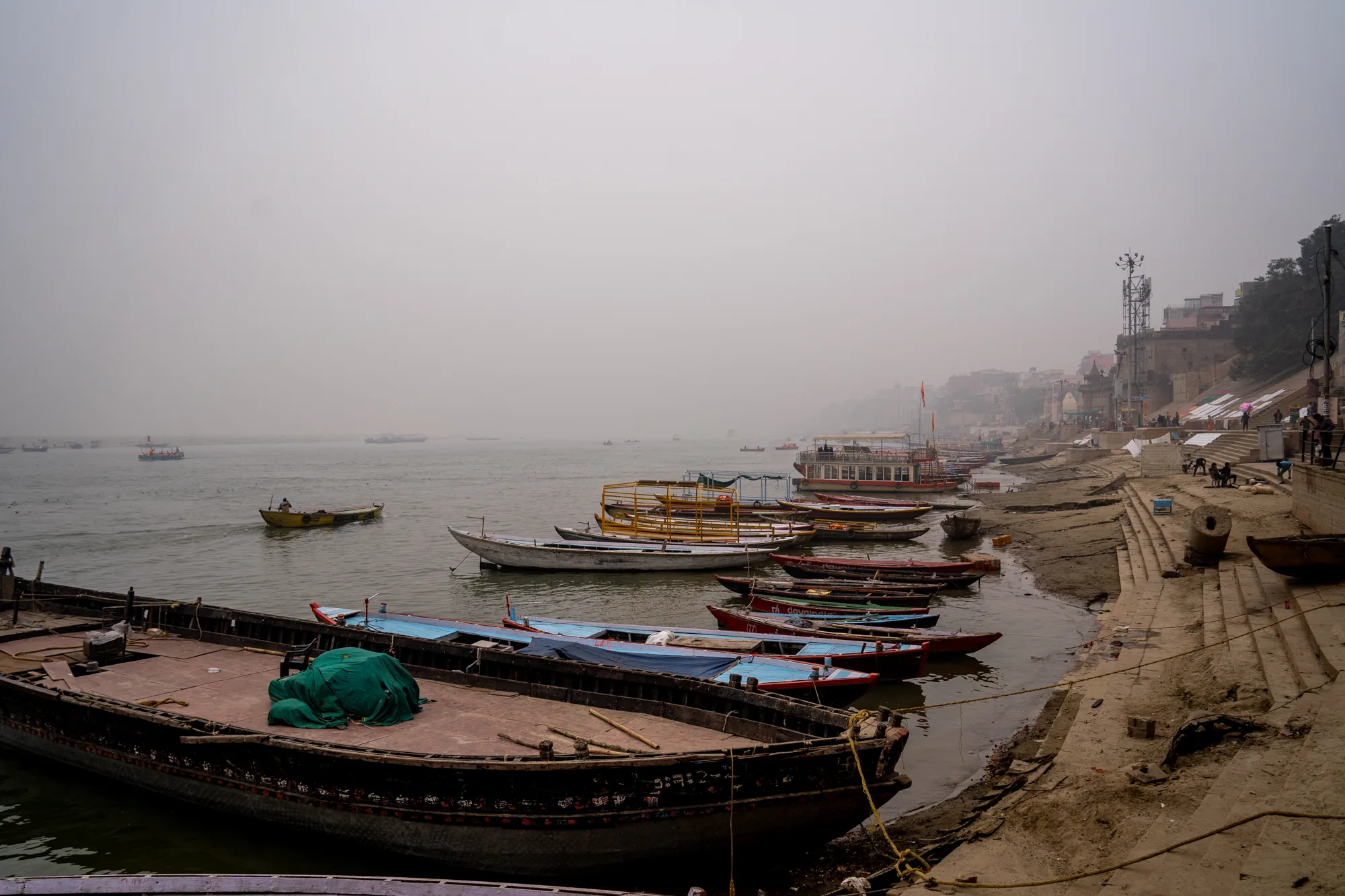 写真はインドのバラナシで撮影されたもので、霧が立ち込めるガンジス川とその岸辺の様子を捉えています。画面全体は灰色がかった色調で覆われており、視界は非常に悪く、遠景はほとんど見えません。  手前には、老朽化した木造の船が複数停泊しています。船体は様々な色で塗られていますが、経年劣化が進み、塗装が剥がれ落ちている箇所も見られます。手前の大きな船には緑色のシートがかけられています。  川面には、小さな手漕ぎボートがいくつか浮かんでおり、そのうちの1隻には人が乗っています。遠くには、かすかに他の船の影が見えます。  岸辺は干上がっており、土色の階段が川へと続いています。階段にはロープが張られており、人々が座ったり、立ち話をしたりしている様子が見えます。背景には建物が立ち並び、通信塔が空に向かってそびえ立っています。岸辺の建物群や階段にいる人々の様子から、この場所が生活の一部として利用されていることが伺えます。全体的に静かで、少し寂しげな雰囲気が漂っています。