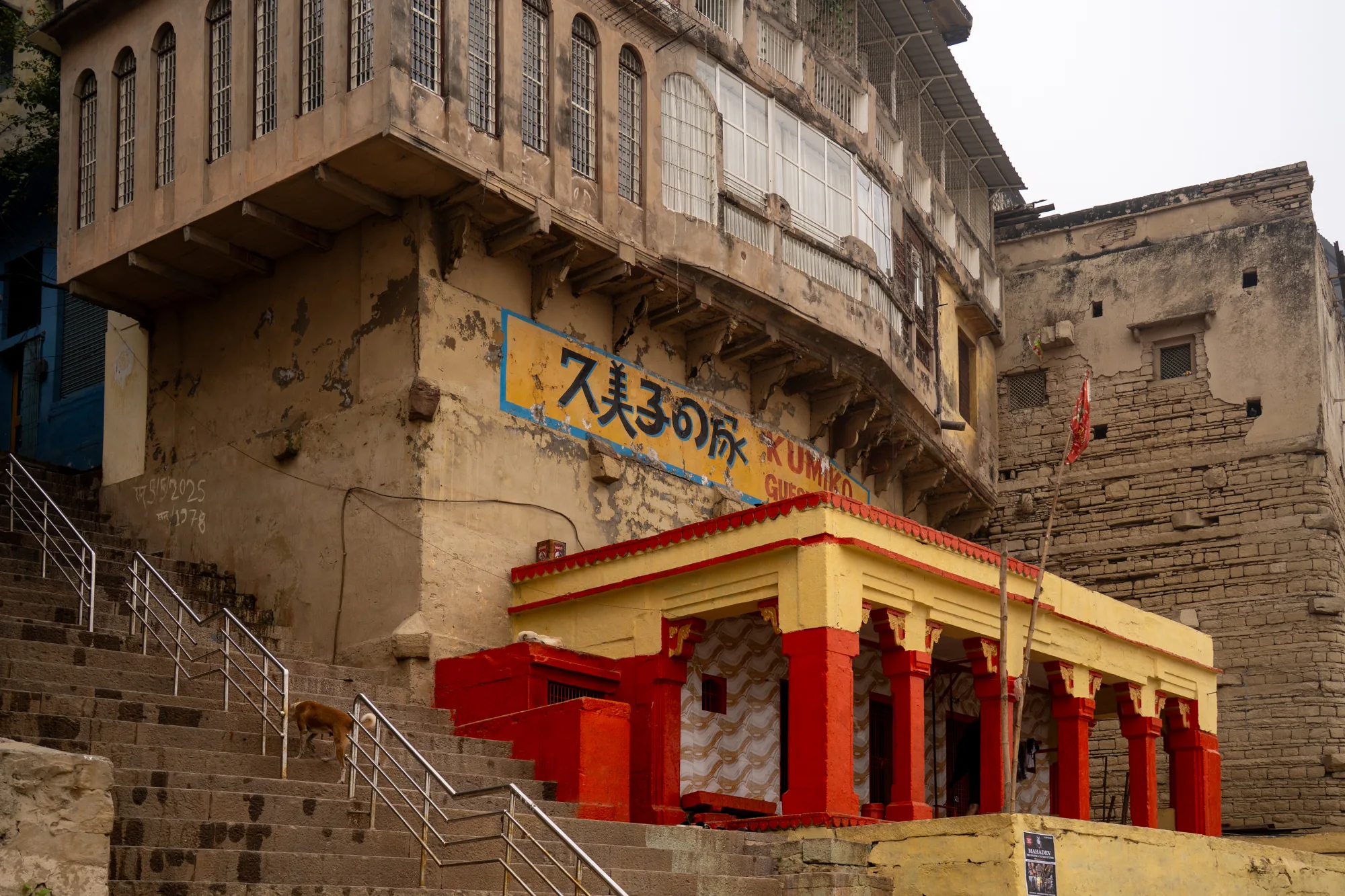 写真はインドのヴァラナシで撮影されたものと思われます。石造りの階段があり、手すりがついています。階段を上がった先に、赤と黄色のコントラストが鮮やかな、柱が並んだ小さな祠のような建物があります。その奥には、古びたベージュ色の建物がそびえ立っています。建物の壁には日本語で「ス美子の床 KUMIKO GUEST」と書かれた看板が掲げられています。建物の外観は経年劣化が進んでおり、剥がれかけた塗装やひび割れが見られます。空は曇り空で、全体的に落ち着いた、歴史を感じさせる雰囲気が漂っています。階段を犬が上がってきています。