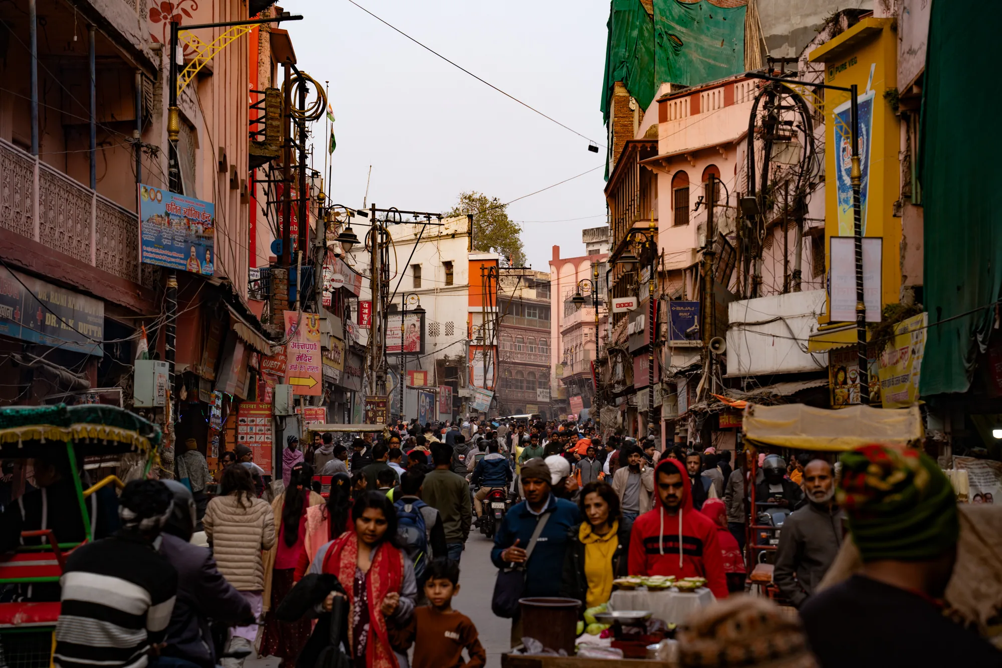 写真はインドのバラナシで撮影されたもので、狭い通りが人で埋め尽くされている様子を捉えています。通りは建物で両側を囲まれ、建物の壁には様々な看板や広告が貼り付けられています。電線が複雑に絡み合い、空を覆っています。通りには、様々な服装の人々が歩いており、歩行者だけでなく、バイクやリクシャーも混在しています。人々の表情は様々で、穏やかな表情の人もいれば、真剣な表情の人もいます。全体的に、活気があり、エネルギーに満ち溢れた雰囲気です。写真の色調はややくすんでおり、埃っぽさや生活感を感じさせます。通りに並んだお店や、行き交う人々の様子から、バラナシの日常風景が伝わってきます。
