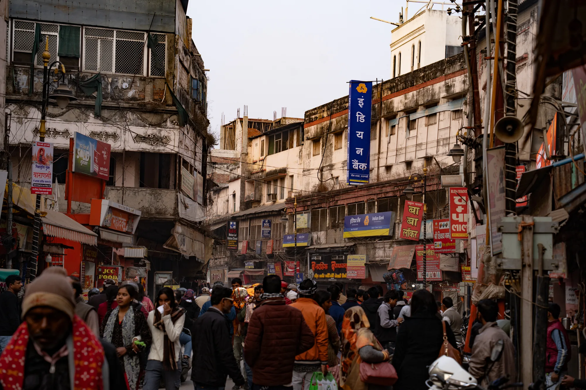 この写真は、インドのヴァラナシで撮影された、混雑した市場の様子を捉えています。狭い通りには多くの人々が行き交い、活気に満ちた雰囲気です。建物の壁には様々な広告や看板が掲げられ、情報が溢れています。建物の多くは古く、壁の塗装が剥がれていたり、ひびが入っていたりするなど、歴史を感じさせます。電線が複雑に張り巡らされており、生活感が溢れています。画面手前には歩行者が多く写っており、奥に向かって人通りが増えている様子がわかります。人々の服装は様々で、伝統的なものから現代的なものまで見られます。遠景には白い建物が見え、空は薄曇りのようです。全体的に、混沌としながらもエネルギーに満ちた、ヴァラナシの日常が伝わってくる写真です。