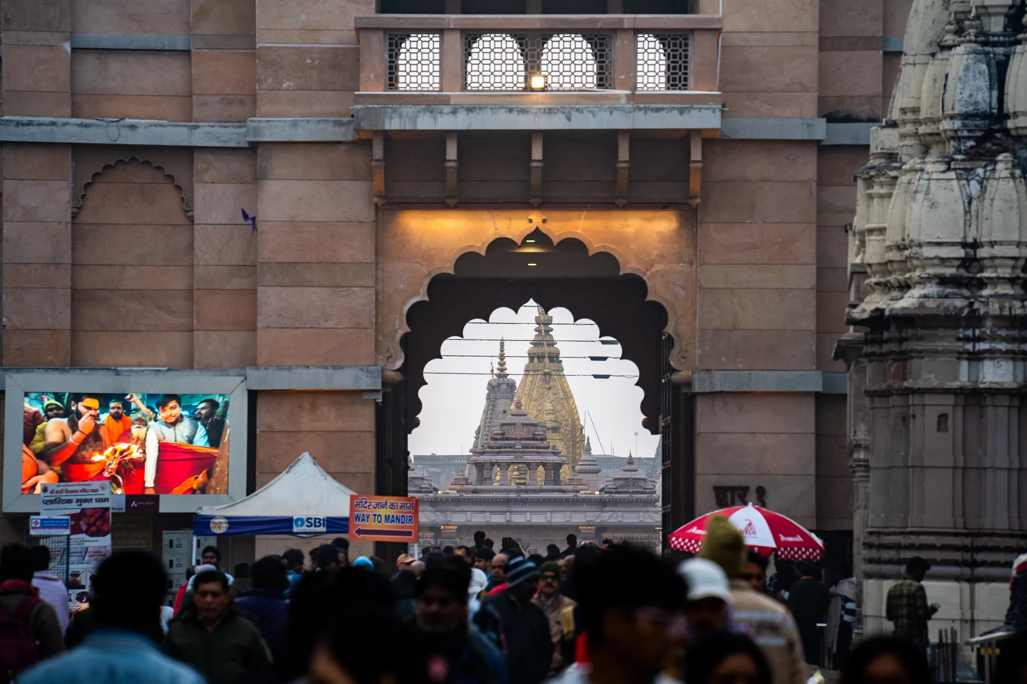 インドのバラナシで撮影された写真です。画面中央には装飾的なアーチ型の門があり、その奥に寺院の尖塔が見えます。寺院は金色に輝いており、神聖な雰囲気を醸し出しています。門の手前には多くの人々が集まっており、寺院へ向かっているようです。画面左側には大きなモニターがあり、何か映像が映し出されています。また、屋台のようなものも見えます。全体的に人々の熱気と信仰心が感じられる写真です。