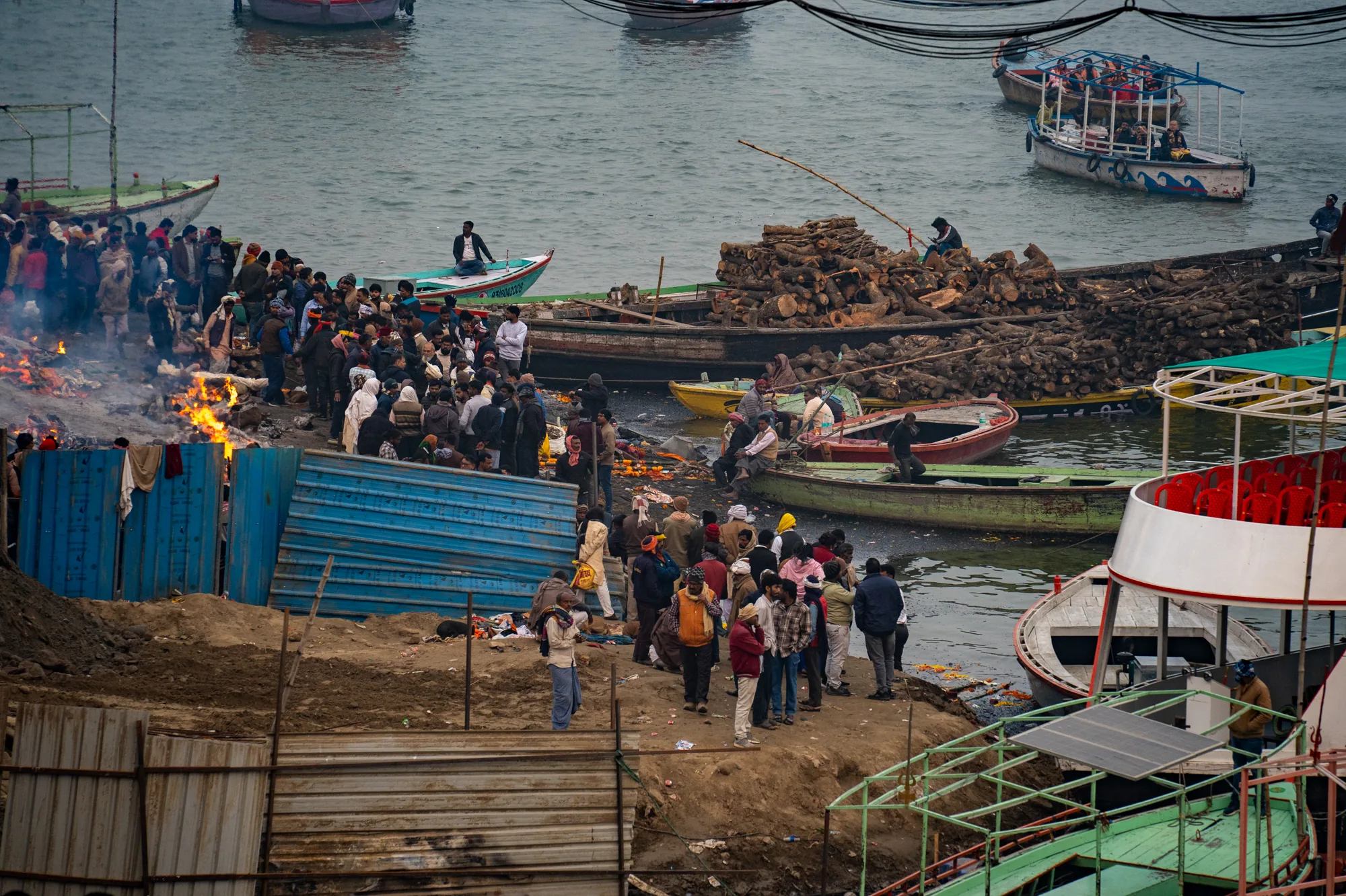 インドのバラナシで撮影された写真です。画面奥にはガンジス川が広がっており、手前は川岸となっています。川岸には多くの人々が集まり、火葬の炎が激しく燃え上がっています。人々は火葬の様子を静かに見守っているようです。川には多くのボートが浮かんでおり、一部には観光客が乗船している様子も見えます。火葬場の周辺には、積み上げられた薪や、様々な種類のボートが雑然と置かれています。写真全体から、生と死が隣り合わせに存在するインドの宗教的な風景が伝わってきます。また、川岸の土や、周囲に散らばるゴミなどから、生活感も感じられます。