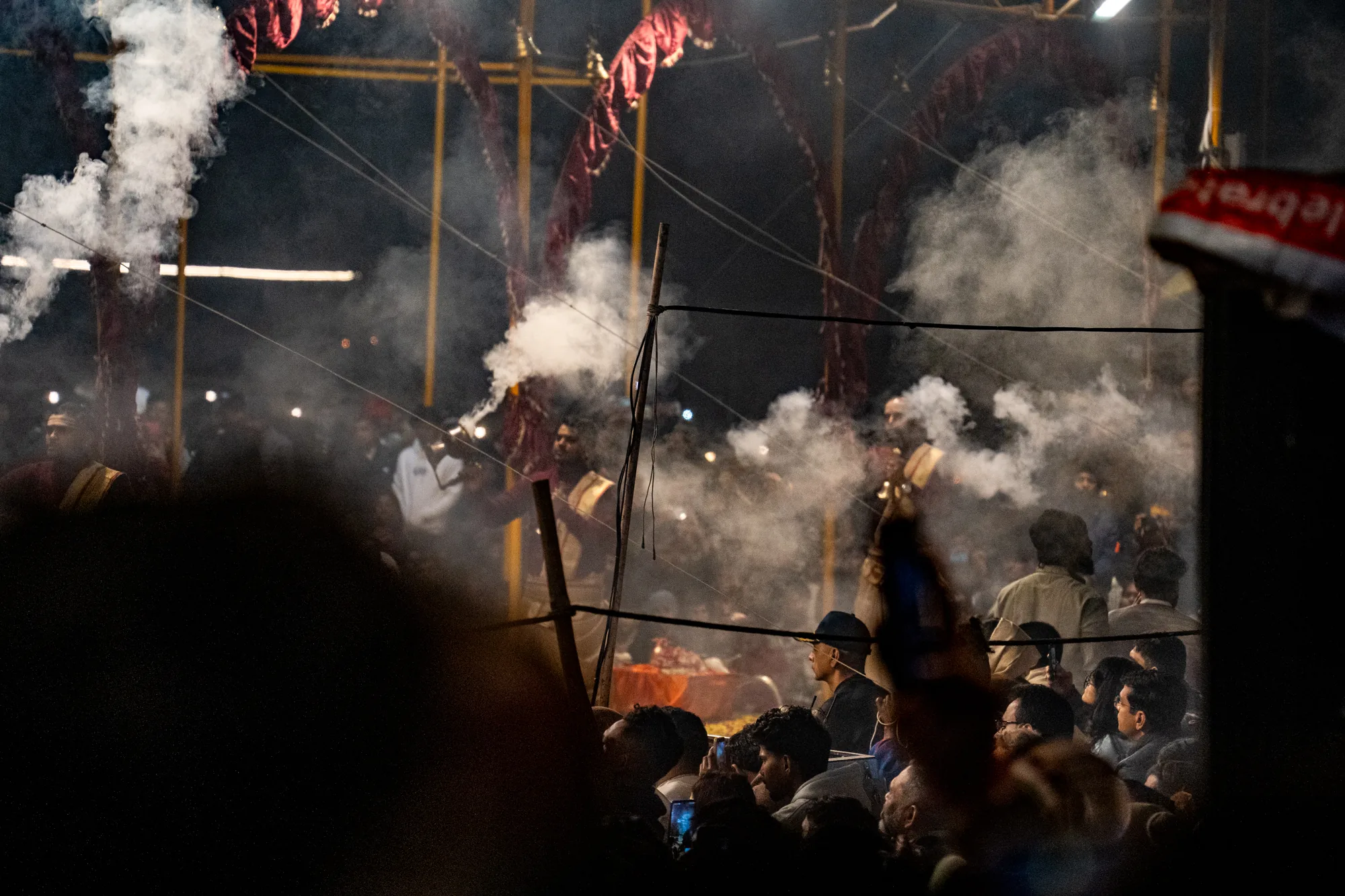 この写真は、インドのバラナシで撮影された宗教的な儀式の様子を捉えています。全体的に暗く、煙が立ち込める中で、人々が集まっている様子がわかります。  手前には群衆の頭部がシルエットで写っており、一部の人はスマートフォンで撮影しているようです。中央には、オレンジ色の布で飾られた構造物が見え、その周りには白い煙が立ち込めています。煙の奥には、儀式を行っていると思われる人々の姿がぼんやりと見えます。彼らは伝統的な衣装を身につけ、楽器を演奏しているようです。  写真全体から、宗教的な熱気と神秘的な雰囲気が伝わってきます。煙と暗闇が、その雰囲気をさらに強調しています。
