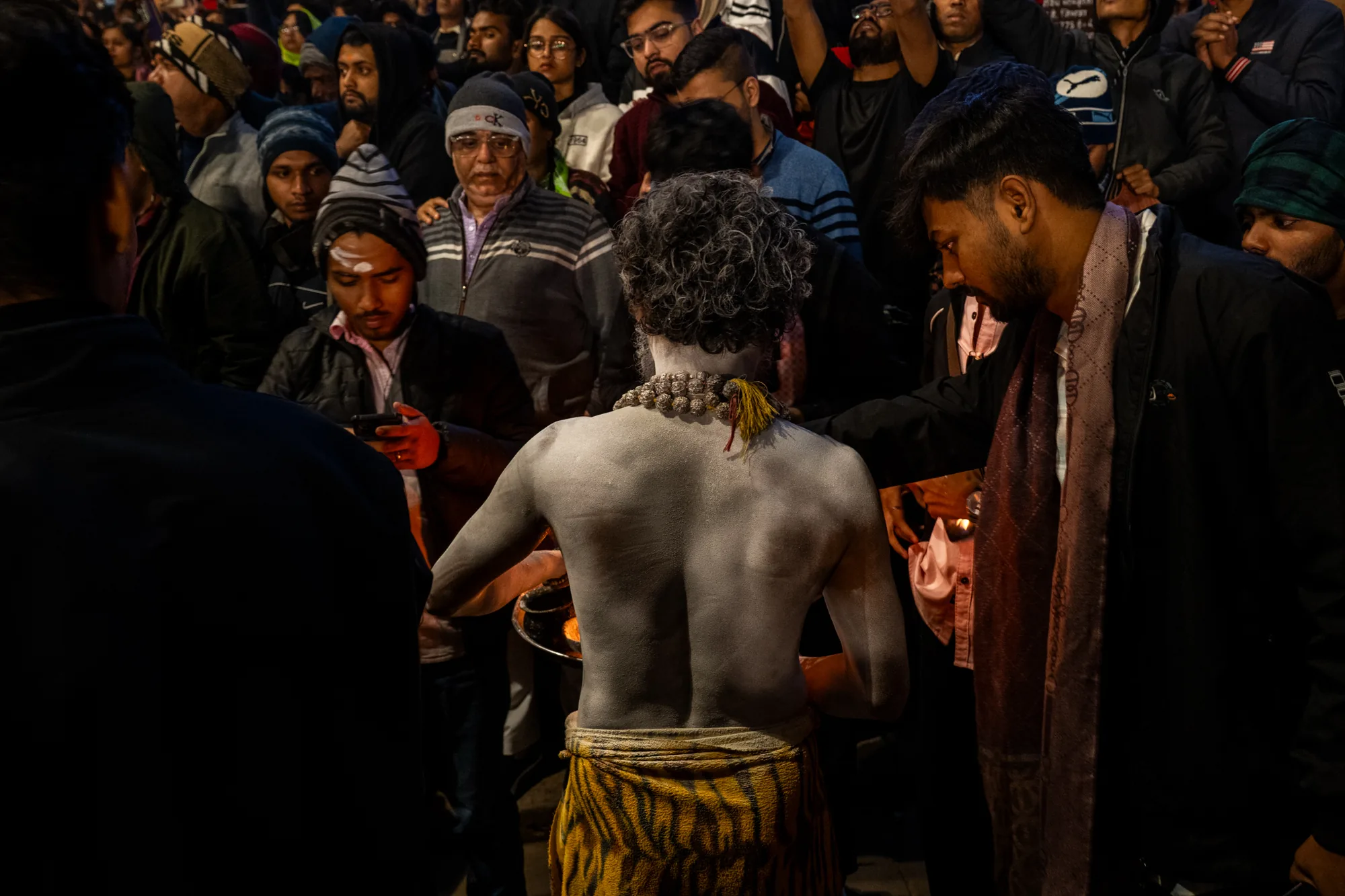 写真はインドのバラナシで撮影されたもので、宗教的な儀式の様子を捉えています。中央に、上半身に白い塗料を塗った男性がおり、虎柄の腰布を身につけています。彼は群衆に背を向け、何かを捧げているように見えます。彼の首には装飾的なネックレスがかけられています。周りには多くの人々が集まっており、様々な服装をしています。一部の人は冬の帽子をかぶり、ジャケットを着ています。群衆の中には携帯電話で何かを見ている人もいます。全体的に、この写真はバラナシの活気に満ちた宗教文化と、儀式に対する人々の熱意を伝えています。照明はやや暗めで、儀式の神秘的な雰囲気を強調しています。