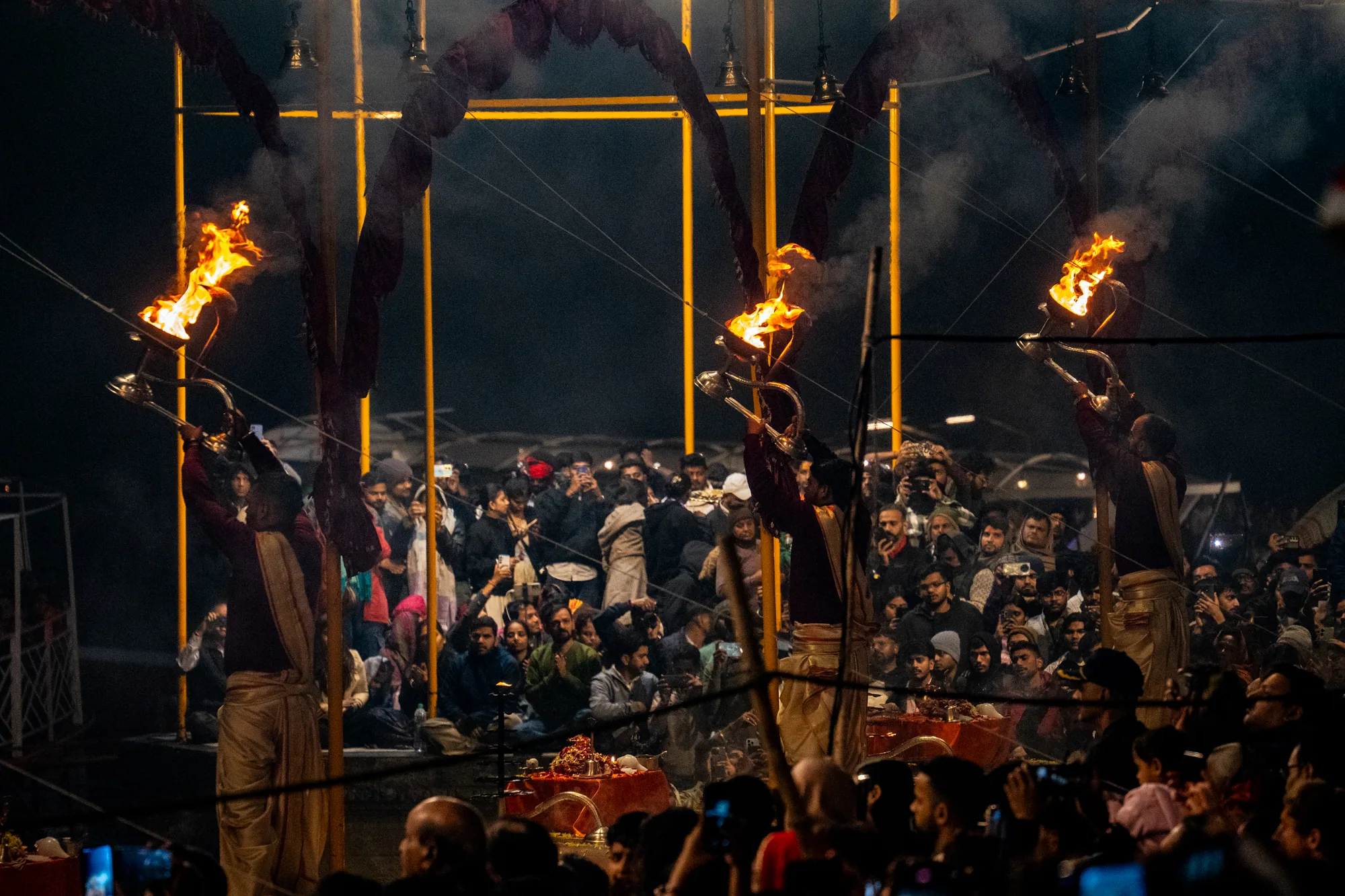 インドのバラナシで撮影された写真です。夜の暗闇の中、多数の人が集まり、宗教的な儀式を観ています。手前には、聖職者と思われる人物たちが３人並び、火のついた容器を持ち上げています。彼らは伝統的な衣装を身に着けており、儀式の厳かさを感じさせます。炎は大きく燃え上がり、周囲に煙が立ち込めています。聖職者の背後には大勢の人がひしめき合っており、スマートフォンで写真を撮ったり、熱心に見入ったりしています。背景には、儀式用の構造物が見え、周囲は暗く、炎の光が際立っています。全体的に神秘的で神聖な雰囲気が漂い、独特の世界観を感じさせる写真です。