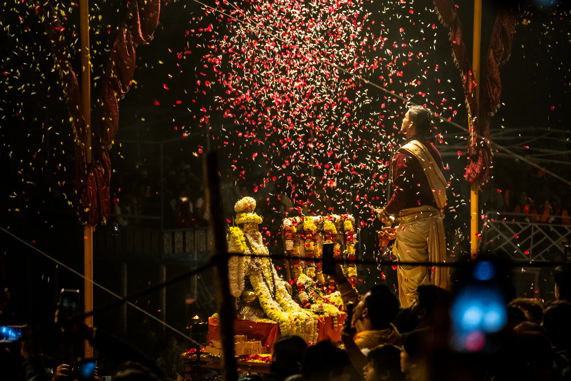 この写真は、インドのバラナシで撮影されたものです。夜の風景の中で、祭りの様子が捉えられています。中央には、花で飾られた像が置かれており、その周りには多くの人々が集まっています。像に花びらを散らしている男性がおり、空には赤と白の花びらが舞い散り、幻想的な雰囲気を醸し出しています。群衆の中には、スマートフォンでその様子を撮影している人も見られます。背景は暗く、花びらの光と、花で彩られた像の鮮やかな色彩が際立っています。伝統的な衣装を身に着けた男性や、周囲の人々の熱気が伝わってくるような、活気あふれる一枚です。