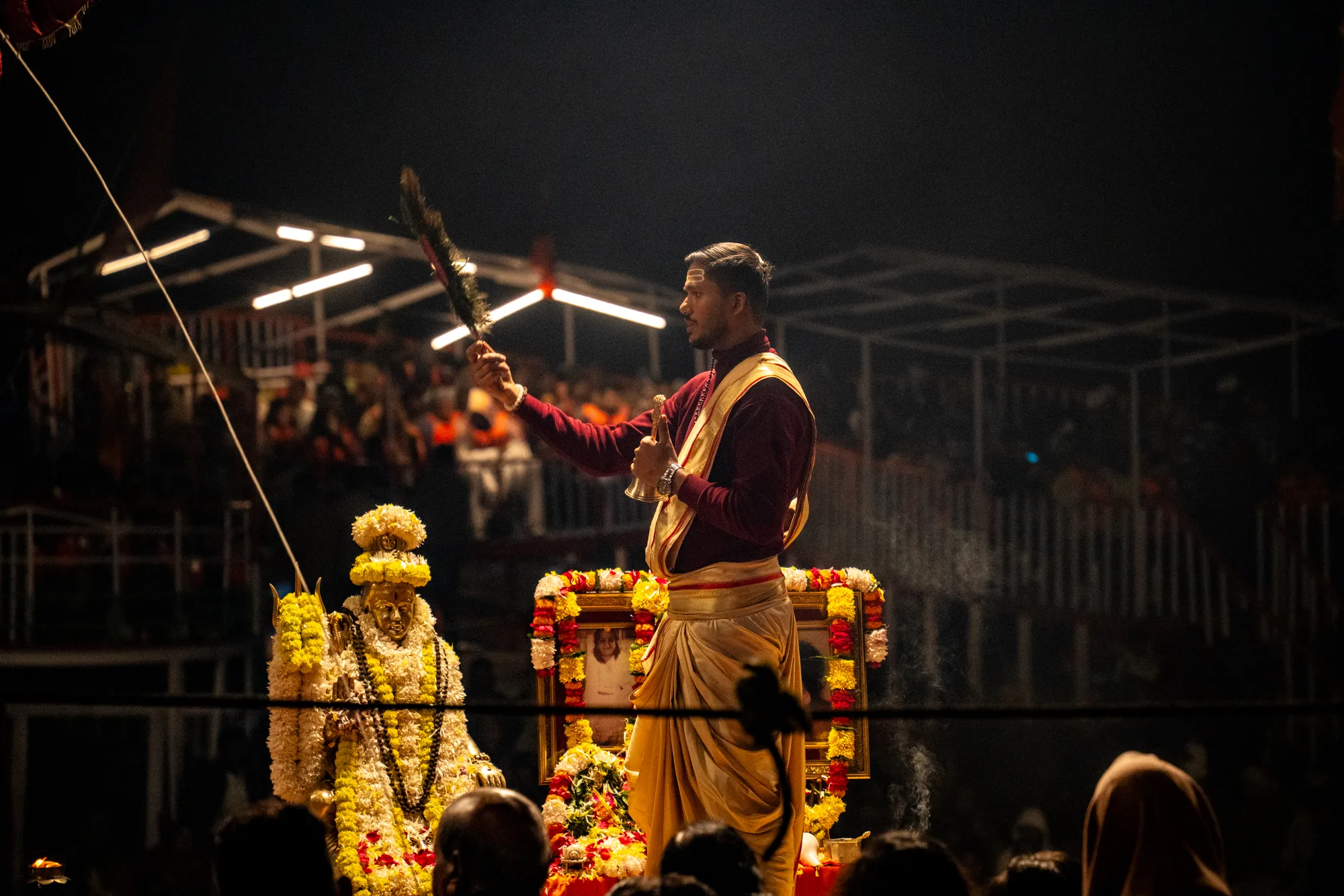 写真はインドのバラナシで撮影されたと思われる、宗教的な儀式の様子を捉えたものです。夜間の暗い背景の中、儀式を行う男性が中心に配置されています。男性は赤紫色のシャツに金色の帯を巻いた衣装を身につけ、右手に装飾されたハエ叩きのようなもの、左手にベルを持っています。彼は真剣な表情で、儀式に集中している様子です。  男性の背後には、花で飾られた祭壇のようなものがあり、その奥にはぼやけていますが、多くの人々が集まっているのが見えます。祭壇の左側には、同じく花で飾られた神像が置かれており、厳かな雰囲気を醸し出しています。写真全体は暗く、スポットライトのような光が儀式を行う男性と祭壇を照らしており、宗教的な神聖さと神秘的な雰囲気を強調しています。煙が立ち上っている様子も確認でき、より神秘的な雰囲気を醸し出しています。