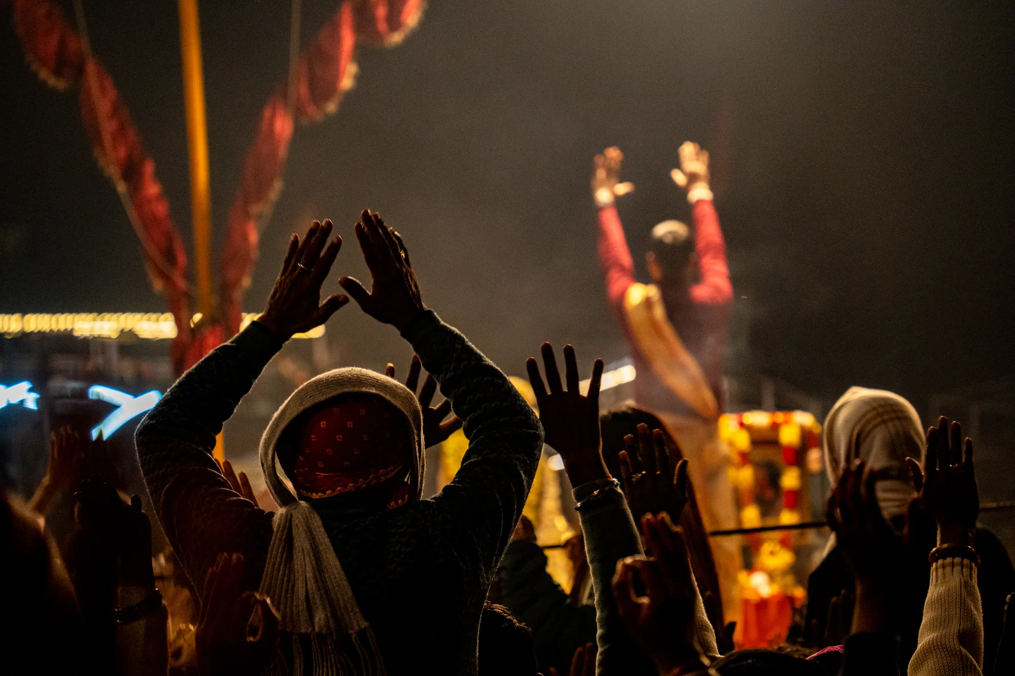 インドのヴァラナシで撮影されたと思われる写真です。多くの人々が手を高く上げ、祈りを捧げている様子が捉えられています。暗い背景の中で、人々のシルエットが浮かび上がり、神秘的な雰囲気を醸し出しています。ライトアップされた装飾が背景にぼんやりと見え、お祭りのような特別な行事であることが伺えます。人々の表情は見えませんが、彼らの動作から信仰心の深さが感じられます。全体的に、宗教的な儀式や祝祭の一場面を切り取った、力強く、そして感動的な写真です。