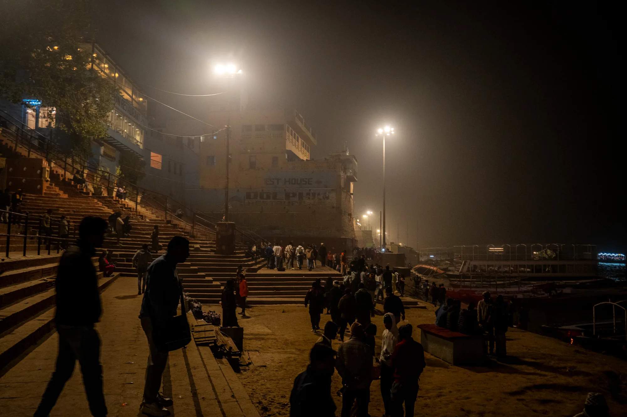インドのバラナシで撮影された写真です。夜の風景で、あたりは深い霧に包まれています。手前の地面には大勢の人が集まっており、それぞれ談笑したり、歩き回ったりしています。シルエットでしか人物の姿は捉えられませんが、活気が伝わってきます。地面の奥には階段があり、階段の上にも人が座っているのが見えます。階段の上には建物があり、建物には「EST HOUSE DOLPHIN RESTAURANT」の文字が見えます。遠くには川のようなものがあり、光が点々と見えます。全体的に暗く、霧がかった雰囲気ですが、街灯の光が暖かく、幻想的な光景を作り出しています。
