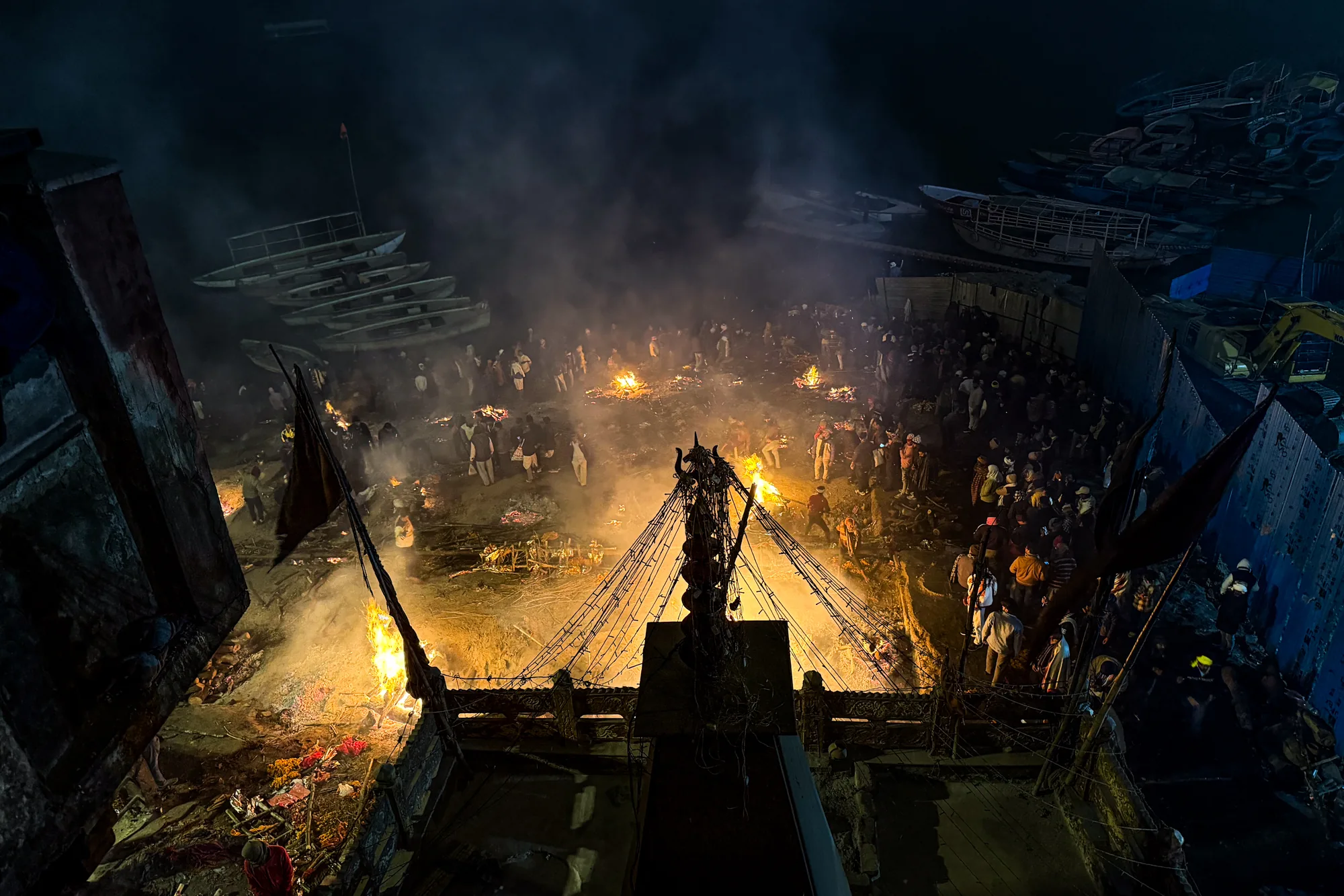 この写真はインドのバラナシで撮影された、夜の火葬場の様子を捉えたものです。高所から見下ろす構図で、画面全体に複数の火が燃え盛っており、煙が立ち込めています。周囲には多くの人々が集まっており、儀式に立ち会っている様子が伺えます。火葬場の近くには、積み重ねられたボートや建設機械も見えます。全体的に暗い色調で、燃え盛る炎が周囲を照らし出し、神聖で厳粛な雰囲気を醸し出しています。写真全体から、生と死が隣り合わせにある場所の独特な雰囲気を感じ取ることができます。