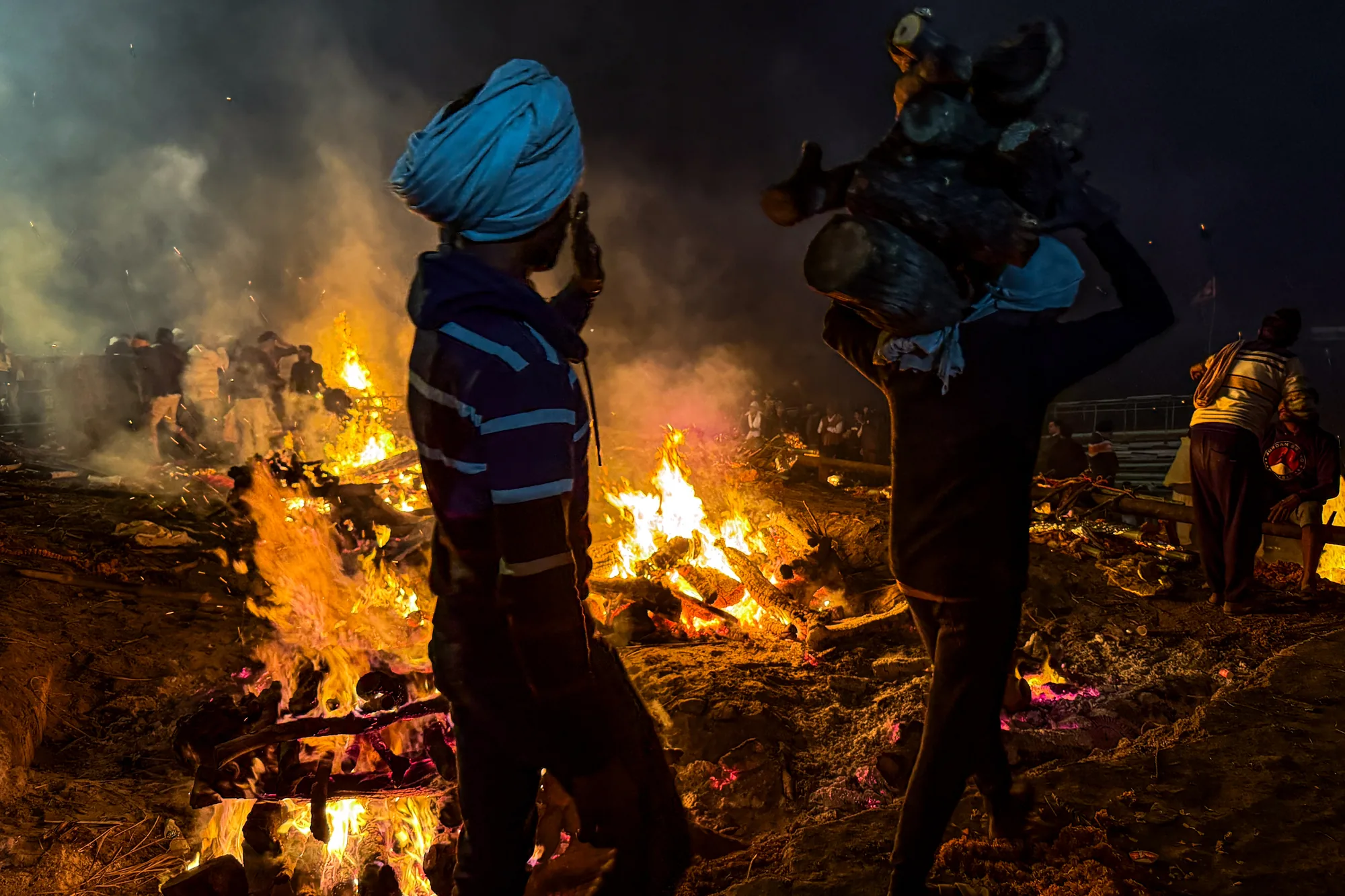 この写真はインドのバラナシで撮影された、夜の火葬場の様子を捉えたものです。画面全体に燃え盛る炎が広がり、周囲は煙に包まれています。炎の前には、ターバンを巻いた男性が立っており、その姿はシルエットとして浮かび上がっています。彼は何かを見つめているように見えます。別の男性は薪を担いで歩いています。奥には群衆が集まっており、火葬を見守っているのかもしれません。炎の色はオレンジ色や赤色、黄色などが混ざり合い、力強く、同時にどこか神秘的な雰囲気を醸し出しています。写真全体を通して、死と再生、そして信仰といったテーマが感じられます。