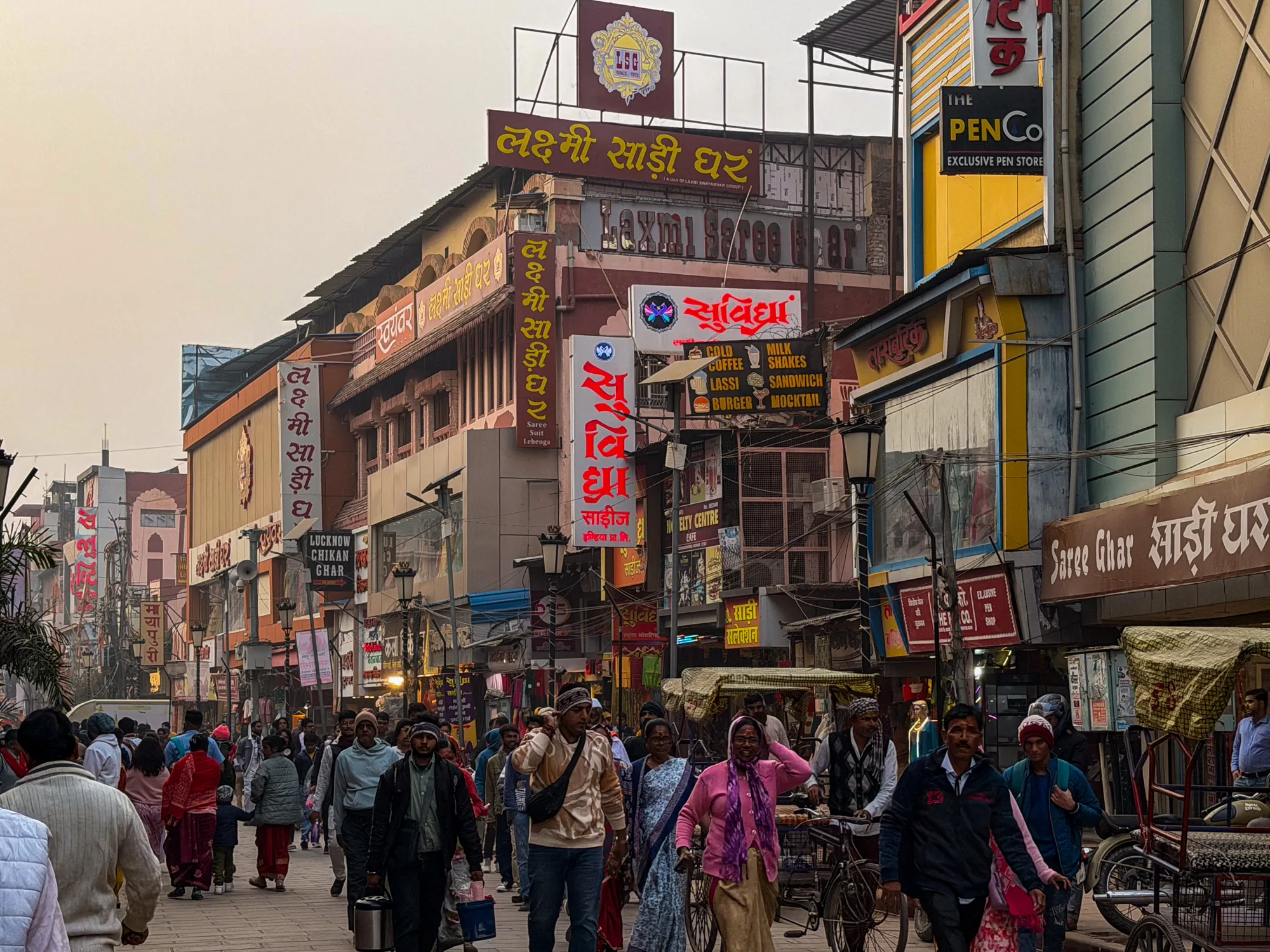 この写真はインドのバラナシで撮影された街並みの様子です。通りには多くの人々が行き交い、活気のある雰囲気が伝わってきます。建物はカラフルな看板で彩られ、「Lakshmi Saree Ghar」や「The Pen Co」といったお店の名前が書かれています。電線が複雑に絡み合い、異国情緒を醸し出しています。通りには歩行者だけでなく、自転車やリクシャーも走っており、生活感にあふれています。全体的に少し霞がかったような空が、落ち着いた印象を与えています。人々の服装も様々で、サリーを着た女性やカジュアルな服装の男性など、多様なファッションが見られます。お店の看板や装飾から、衣料品店や飲食店が多いことが伺えます。