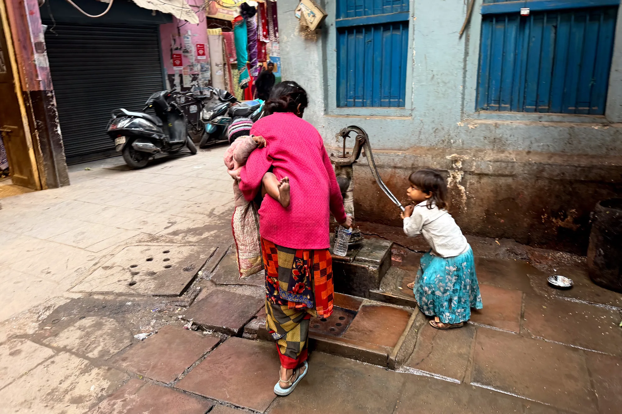 写真はインドのバラナシで撮影されたもので、路地の日常風景を捉えています。中央には、赤ちゃんを抱いた女性が写っています。女性はピンクのニットに、鮮やかな柄のスカートとズボンを重ね着し、サンダルを履いています。そのそばには、水色の花柄のスカートと白いシャツを着た幼い女の子がおり、手押しポンプのレバーを操作しようとしています。壁には青い窓枠の窓があり、壁の色はくすんだ水色です。路地の奥にはオートバイが数台駐車しており、建物にはポスターのようなものが貼られています。路面はタイル張りですが、水で濡れており、生活感が出ています。全体的に、インドの日常的な風景を写し出したスナップショットという印象です。