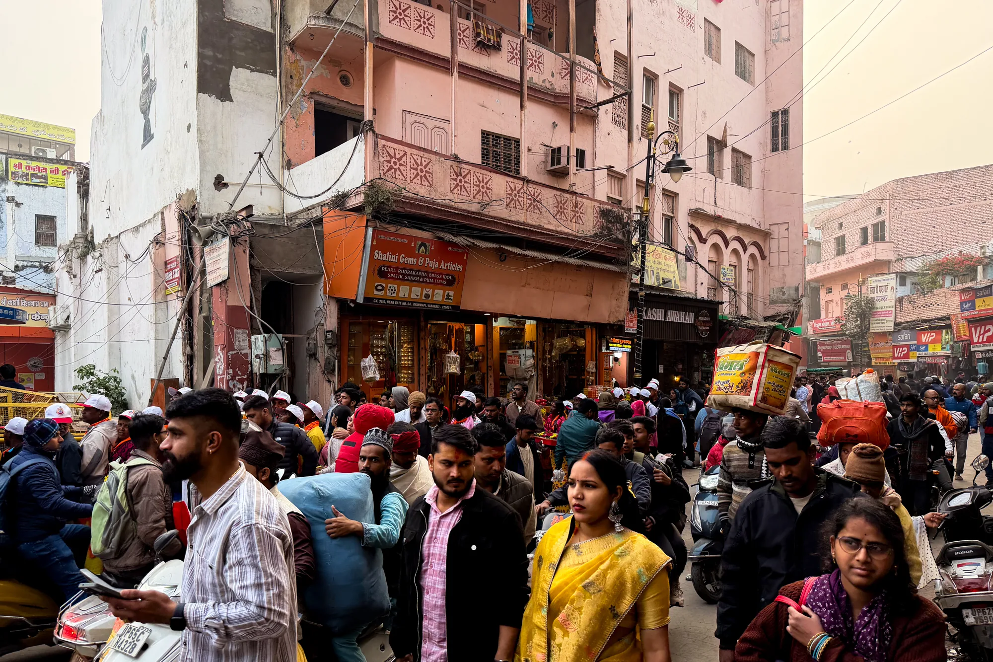 写真はインドのバラナシで撮影された、活気あふれる街の様子を捉えています。狭い路地には多くの人々が行き交い、活気に満ちた雰囲気を醸し出しています。  画面手前には、様々な服装をした人々が写っています。鮮やかな黄色のサリーを着た女性、カジュアルなシャツやジャケットを着た男性、そして伝統的な帽子をかぶった人々など、多様な服装が目を引きます。多くの人がカメラの方を見ており、その表情からは生活感が伝わってきます。  路地にはオートバイが何台か停まっており、人々の移動手段として利用されていることがわかります。背景には、老朽化が進んだ建物が立ち並び、歴史を感じさせる趣があります。建物の壁には、看板や広告などが所狭しと掲げられており、街の賑わいを強調しています。店の看板には、宝飾品やプジャ用品を扱っていることが示唆されています。  写真全体を覆うように、埃っぽい空気と穏やかな光が感じられます。人々の表情や服装、街の様子から、バラナシの日常を垣間見ることができる一枚です。