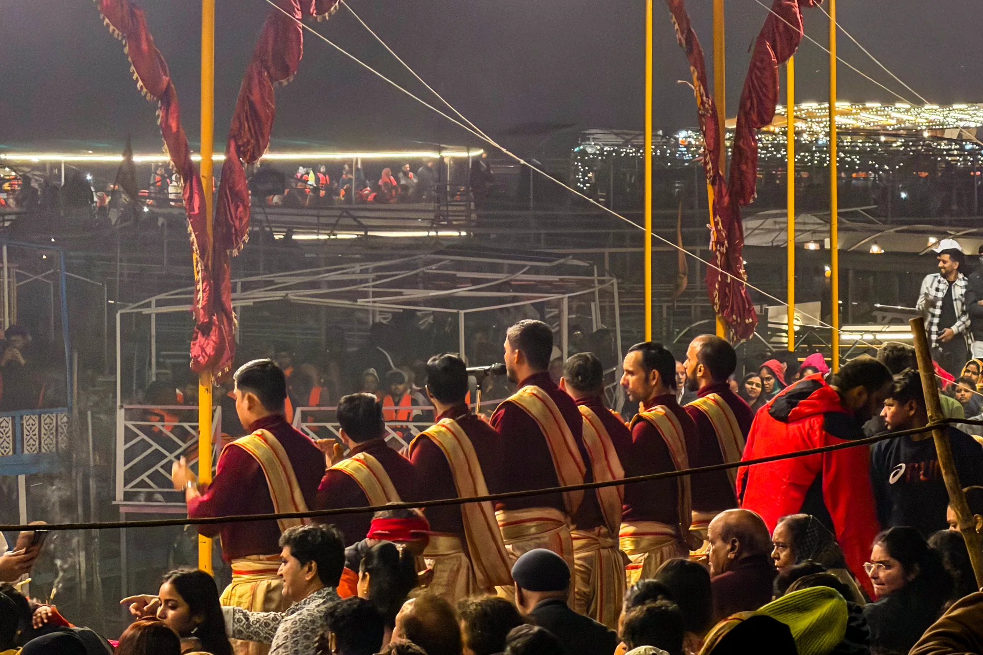 写真はインドのバラナシで撮影された夜の風景を捉えています。手前に並んでいるのは、伝統的な衣装を身につけた複数の男性たちで、彼らは儀式を行っているようです。濃い赤色の服に金の縁取りが施された衣装は、厳かな雰囲気を醸し出しています。彼らの前には多くの人々が集まっており、儀式の様子を見守っている様子が伺えます。群衆の中には、様々な服装をした人がおり、地元の住民から観光客まで様々な人々が集まっていることがわかります。  背景には、ライトアップされた建物や構造物が見え、夜空に浮かび上がるように輝いています。これらの光が、写真全体に暖かく幻想的な雰囲気を加えています。ロープのようなものが画面を横切っており、奥行きを感じさせると同時に、儀式が行われている場所の状況を示唆しています。写真全体を通して、バラナシの宗教的、文化的な深さが感じられます。